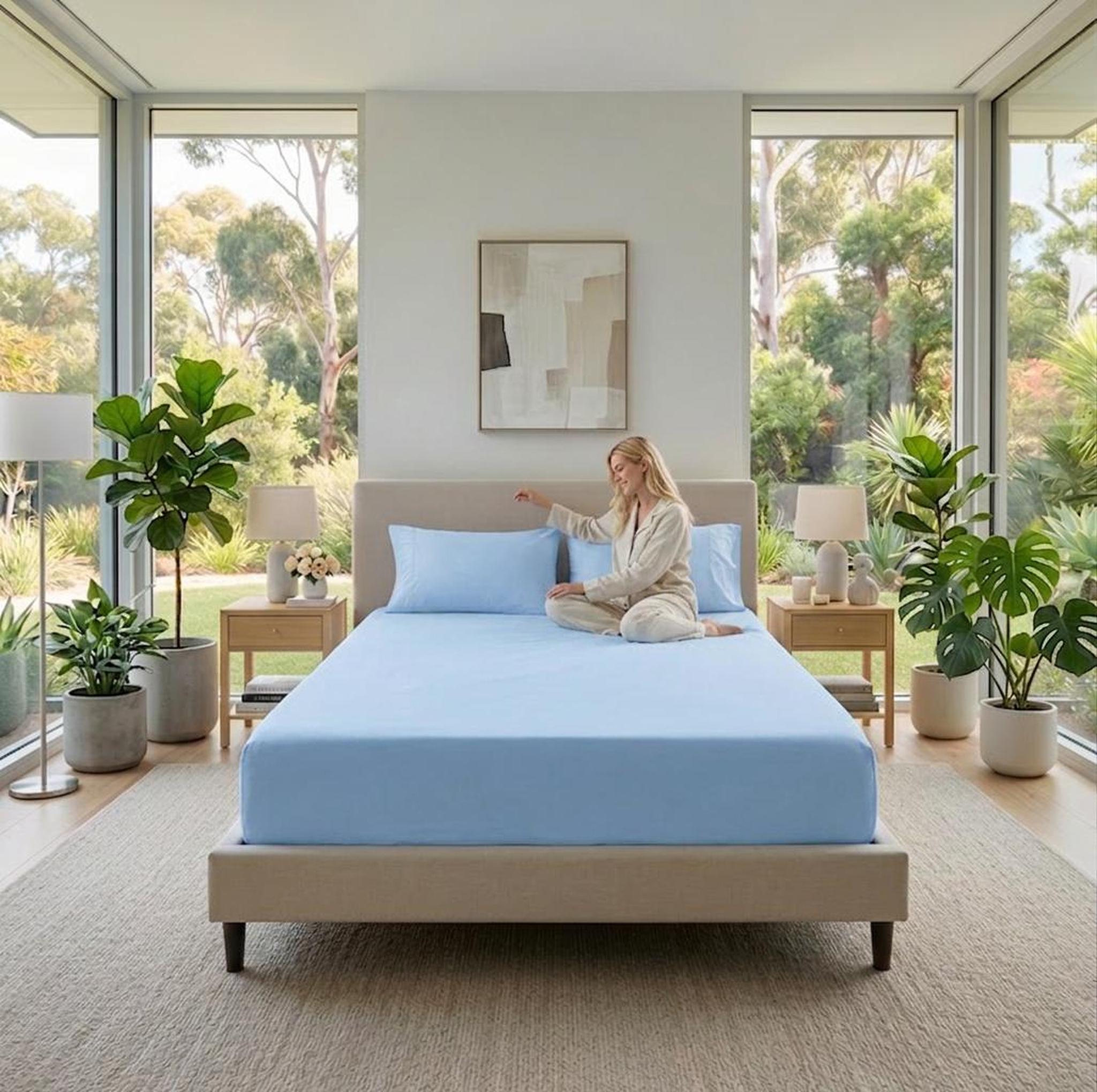 Light blue cotton fitted sheet on a bed with a woman sitting on it, surrounded by plants.