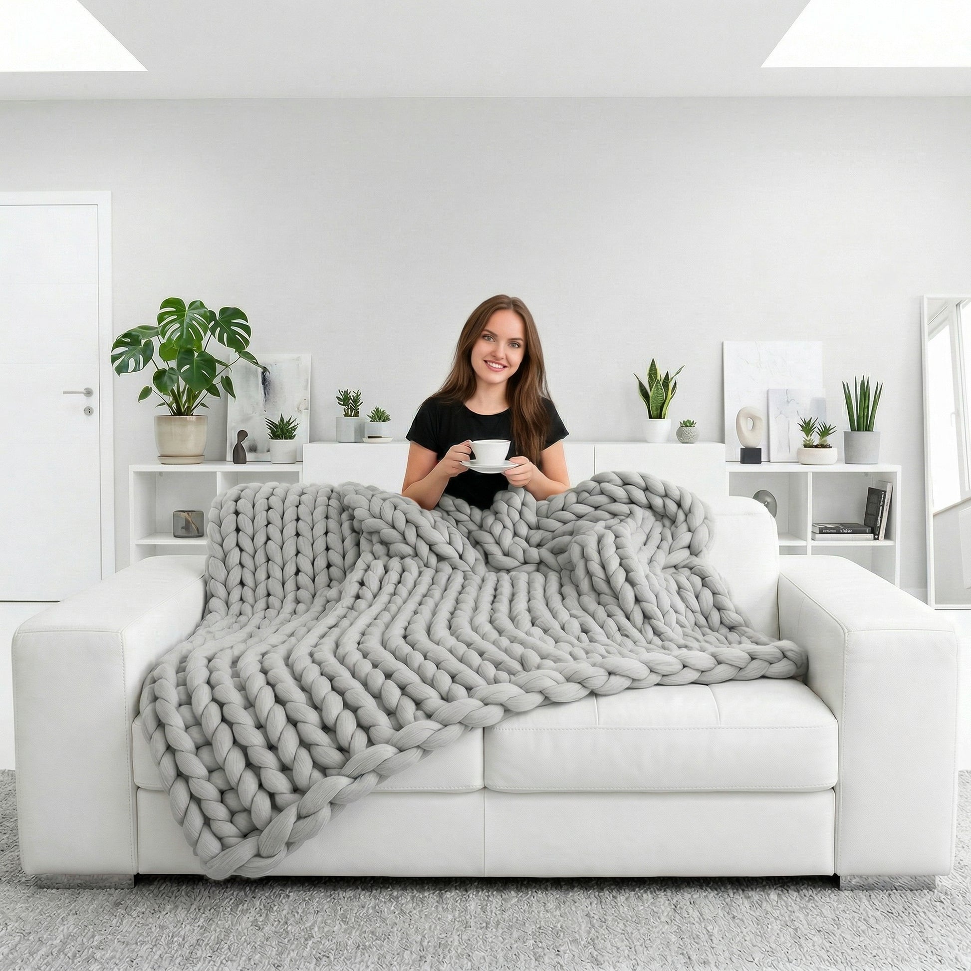 Woman on a white sofa with a large, light gray chunky knit blanket, holding a white cup.