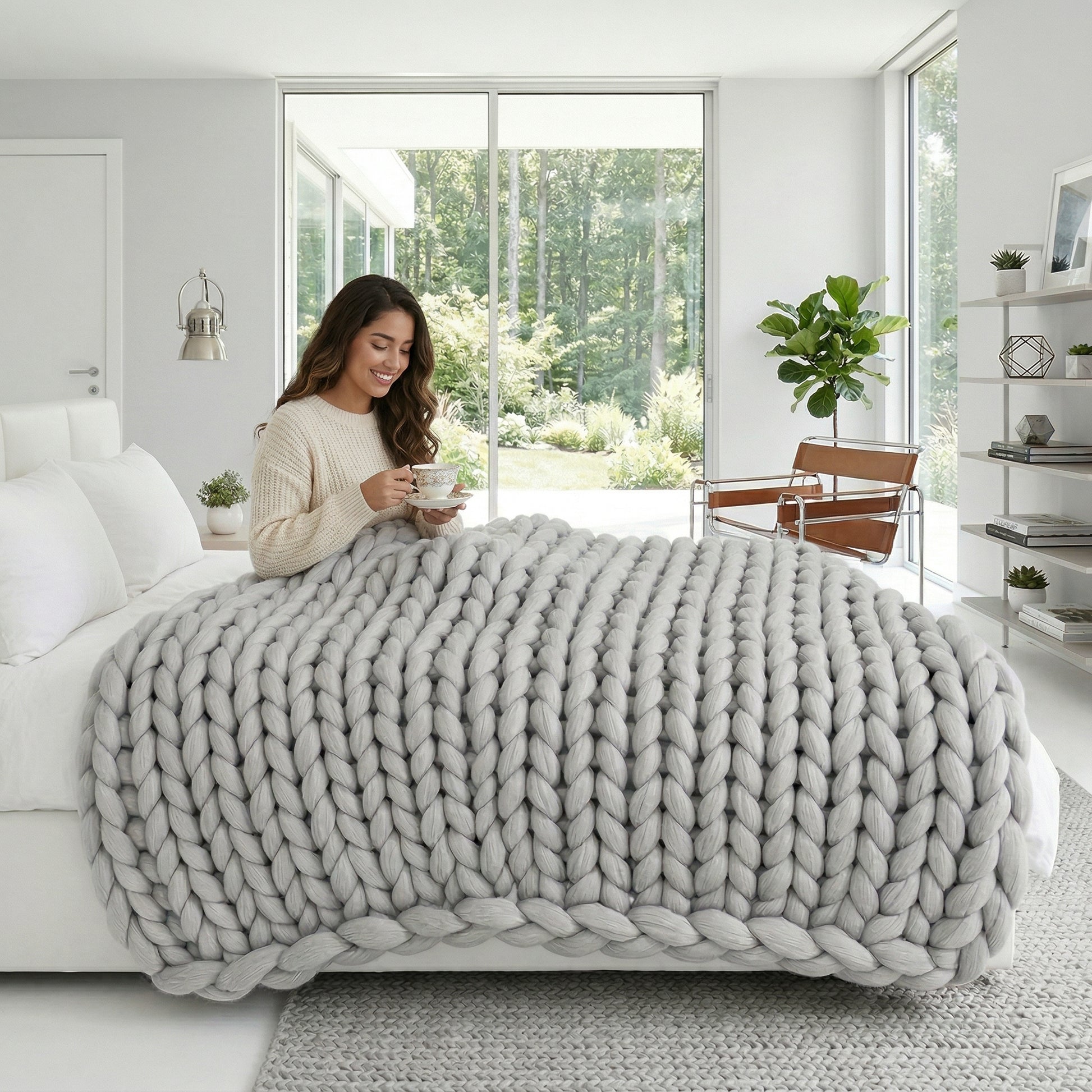 Woman on bed with a large, light grey chunky knit blanket, holding a cup.