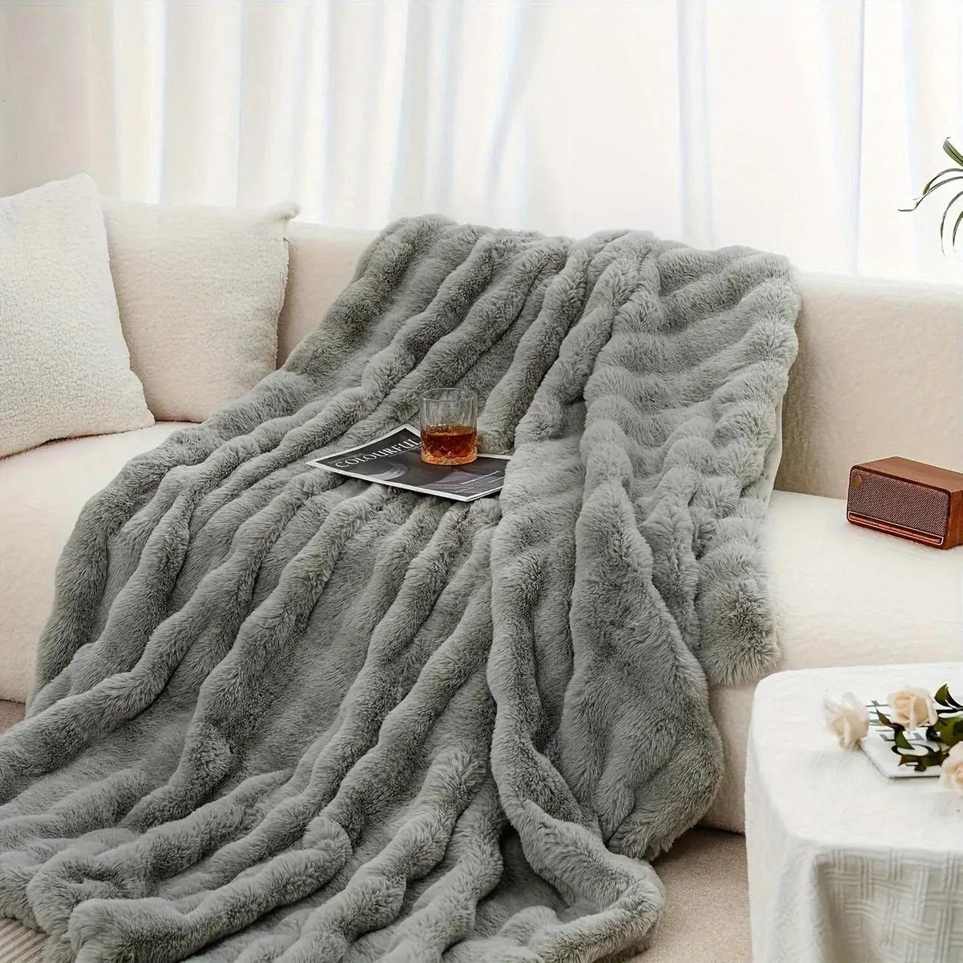 Plush gray faux rabbit fleece throw blanket draped on a white sofa with a drink and magazine.
