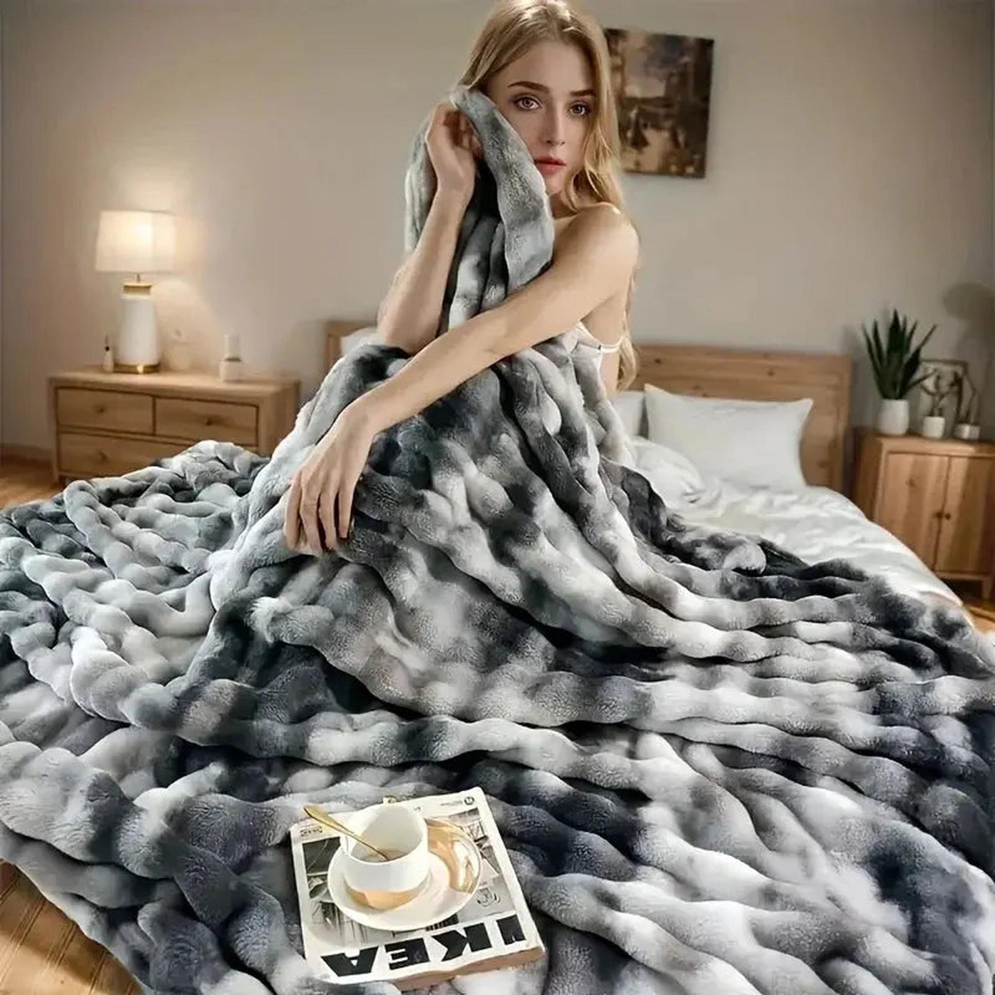 Woman wrapped in a large, plush gray and white faux rabbit fleece throw blanket on a bed.