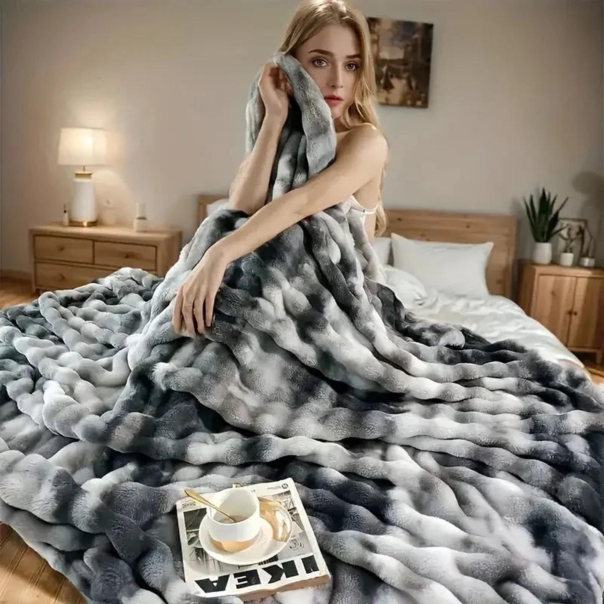 Woman wrapped in a large, plush gray and white faux rabbit fleece throw blanket on a bed.