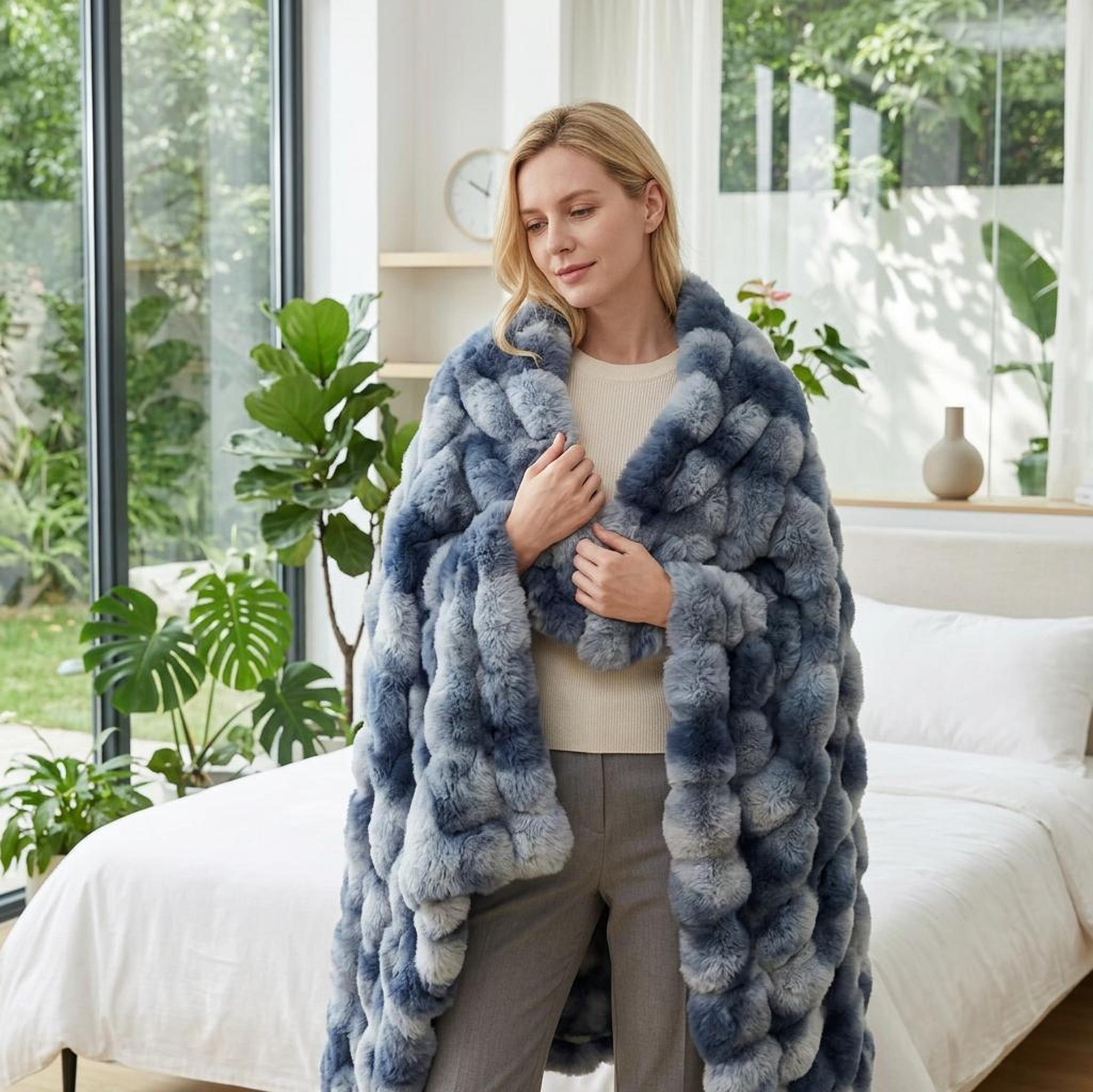 Woman wrapped in a plush blue and grey faux rabbit fleece throw blanket, standing in a bright bedroom.