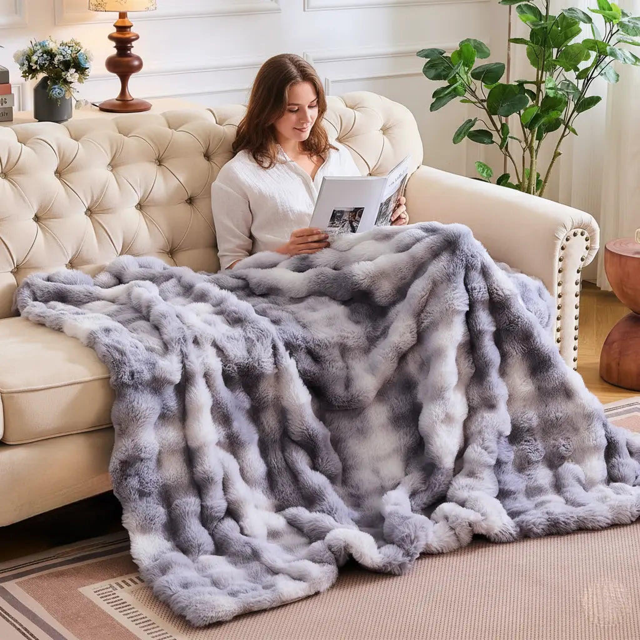 Woman on a cream couch covered by a plush, gray and white faux rabbit fleece throw blanket.