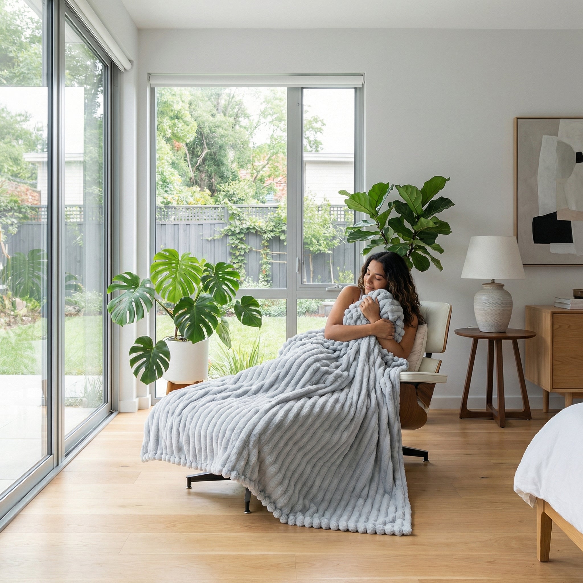 Woman relaxing in a modern living room, wrapped in a soft, light gray CozyNap Flannel Blanket with a ribbed texture.