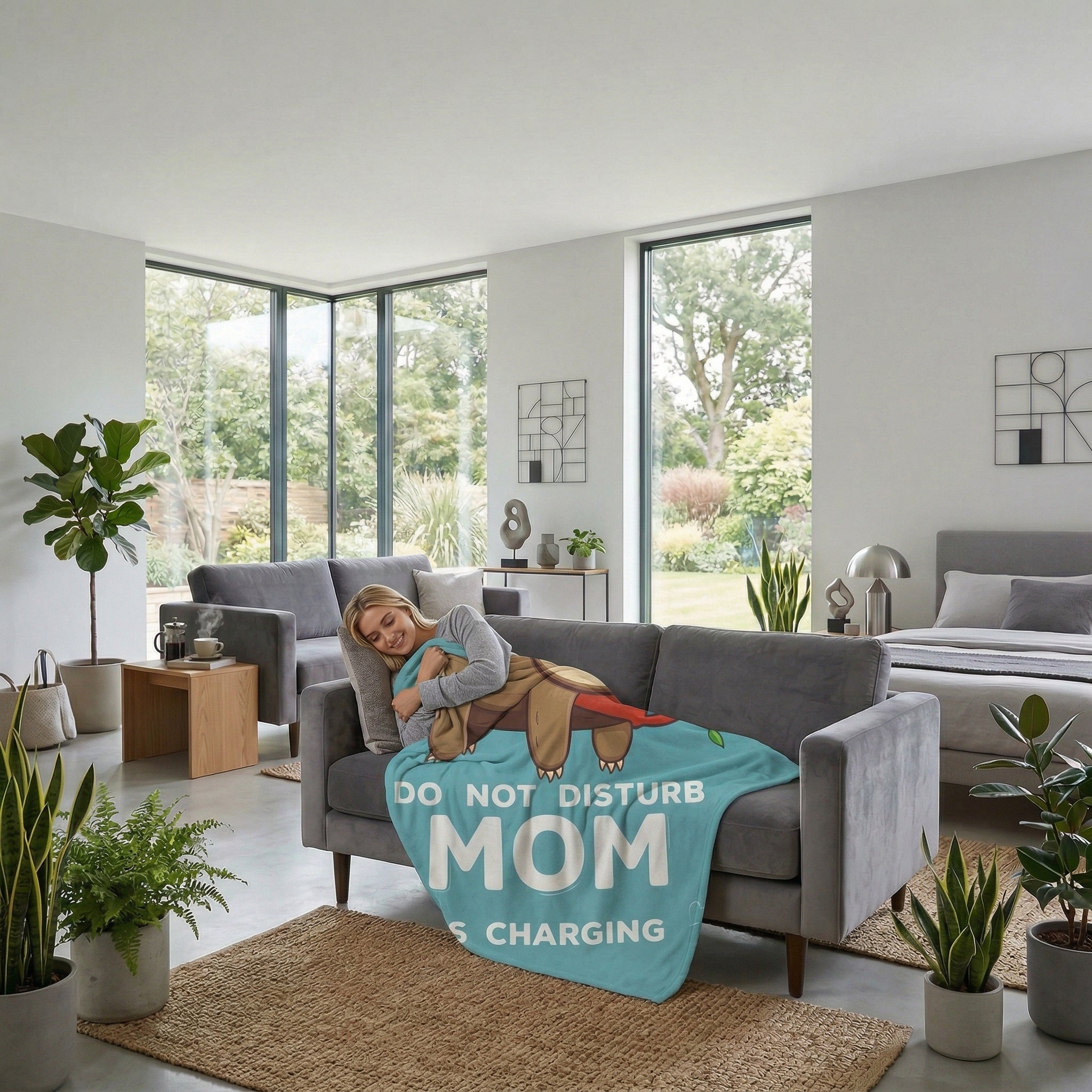 Woman napping on a gray sofa, covered by a light blue "DO NOT DISTURB MOM IS CHARGING" flannel throw blanket.