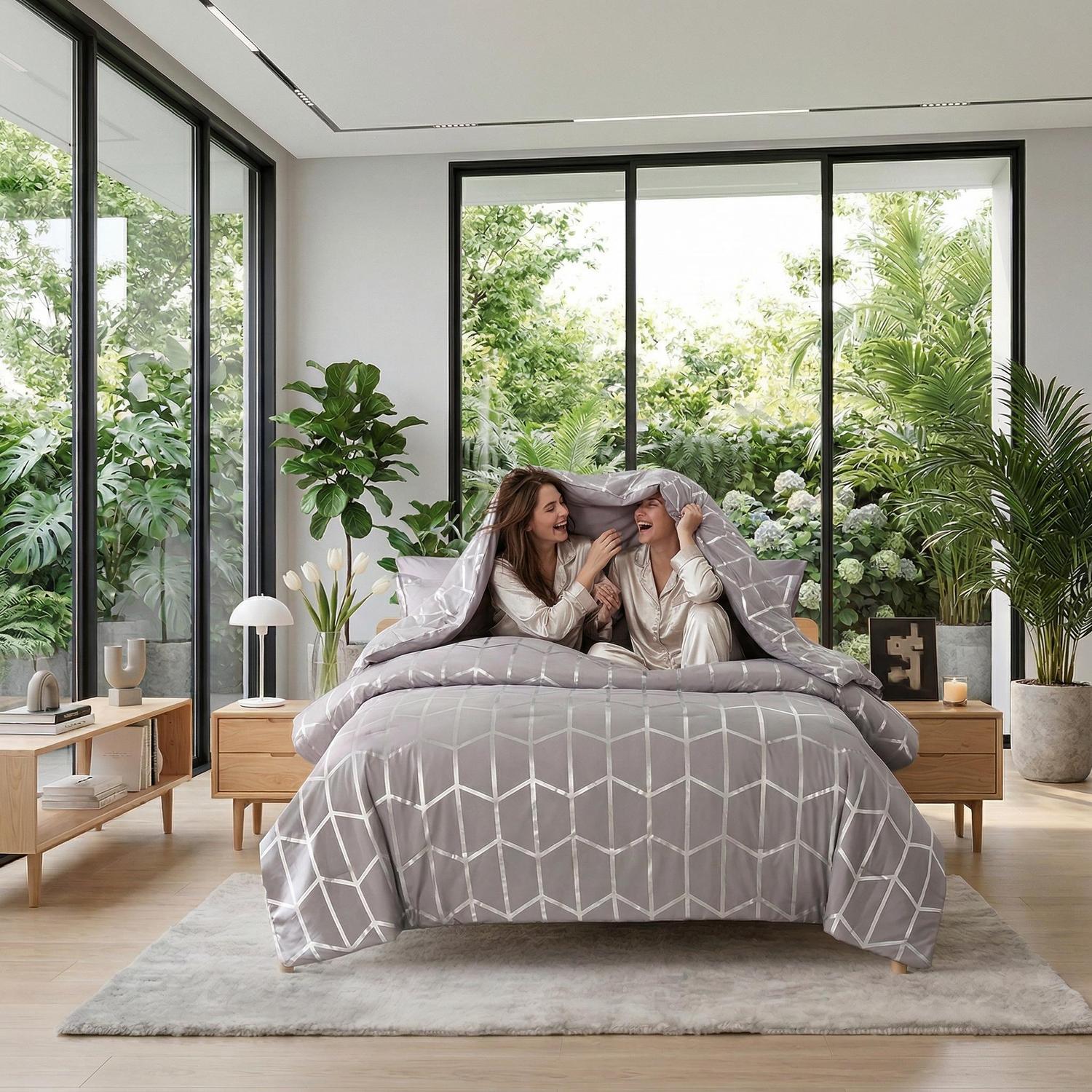 Two women laughing under a grey comforter with a silver geometric foil design in a modern bedroom.