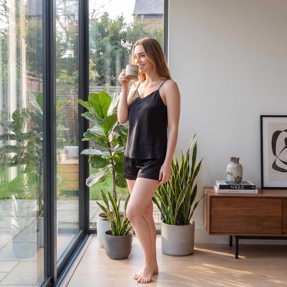 Woman wearing a black silk V-neck camisole and shorts pajama set while holding a mug in a sunlit room.