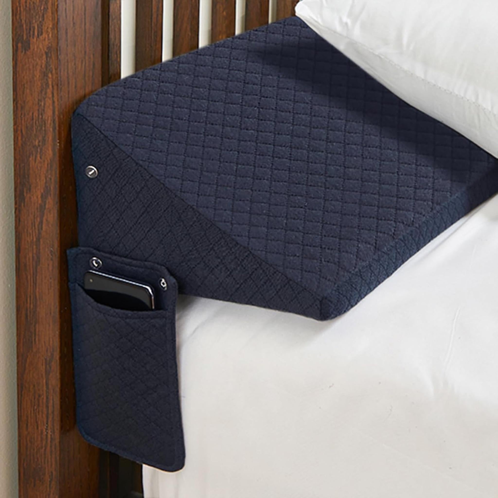 Navy quilted ElevateWedge gap filler pillow with side pocket holding a phone, against a wooden headboard.