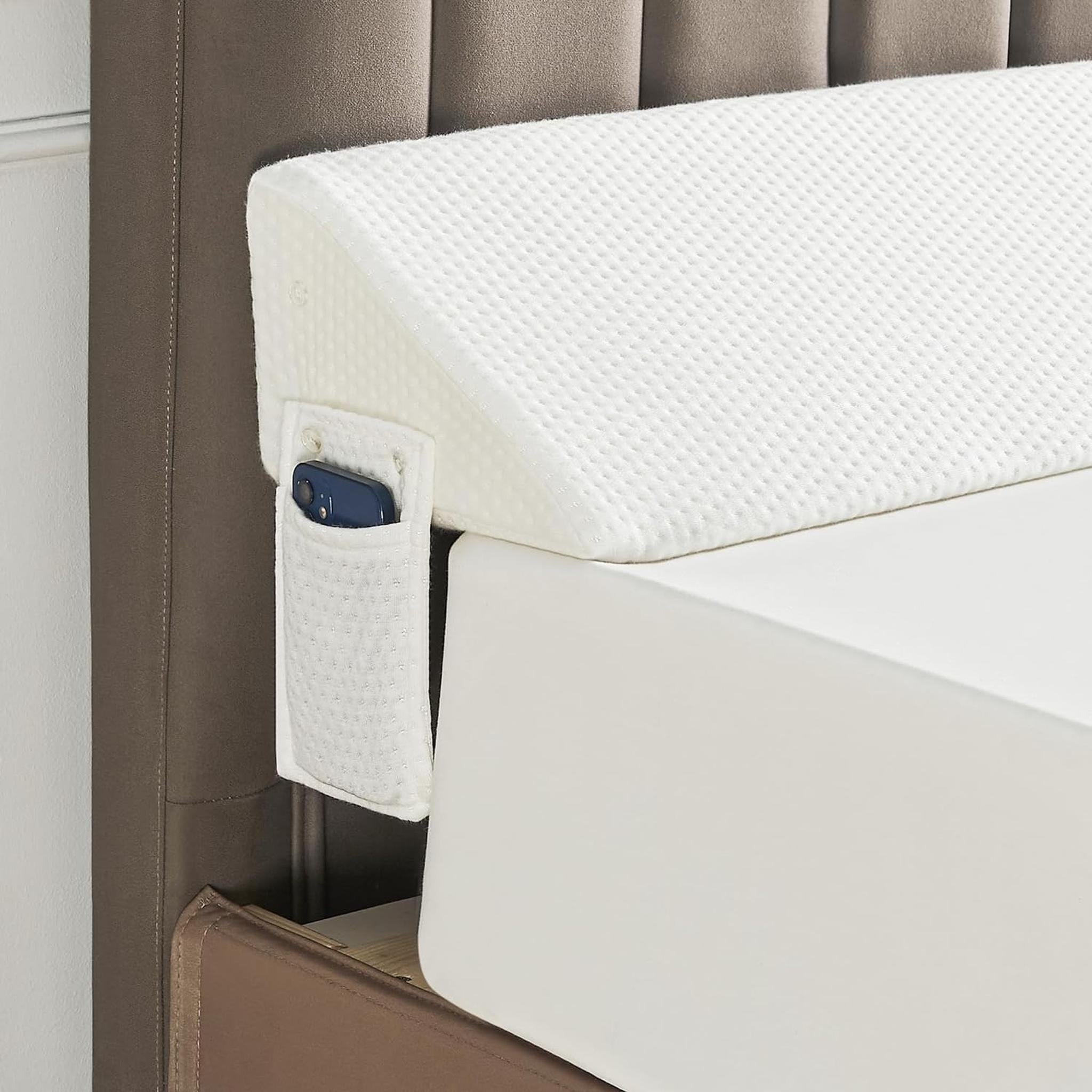 White textured ElevateWedge gap filler pillow with side pocket holding a phone, nestled between a mattress and brown headboard.