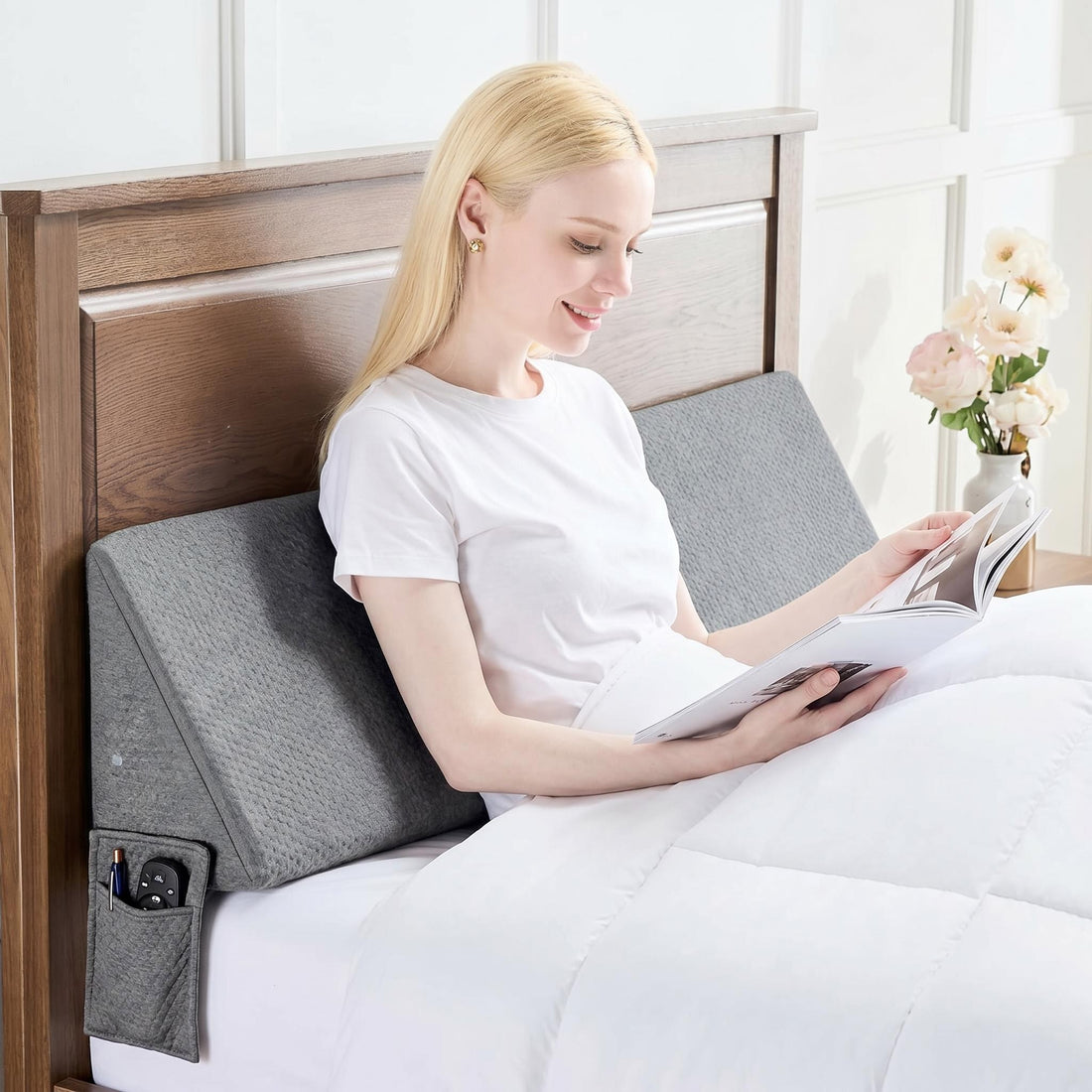 Woman reading in bed with a gray ElevateWedge gap filler pillow, featuring side pockets for remote and phone.