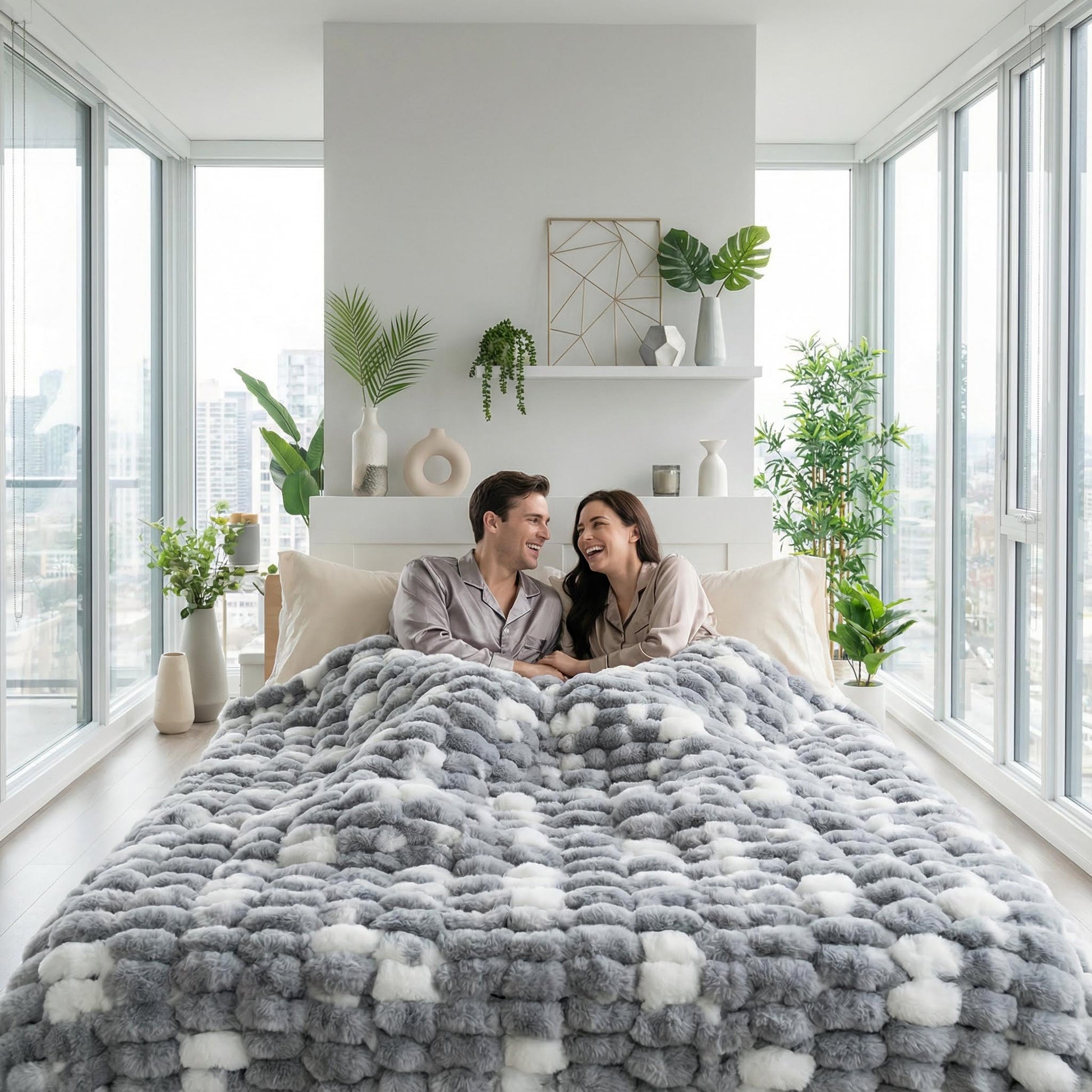 Couple snuggled under a grey and white faux rabbit fur throw blanket on a bed in a bright room.