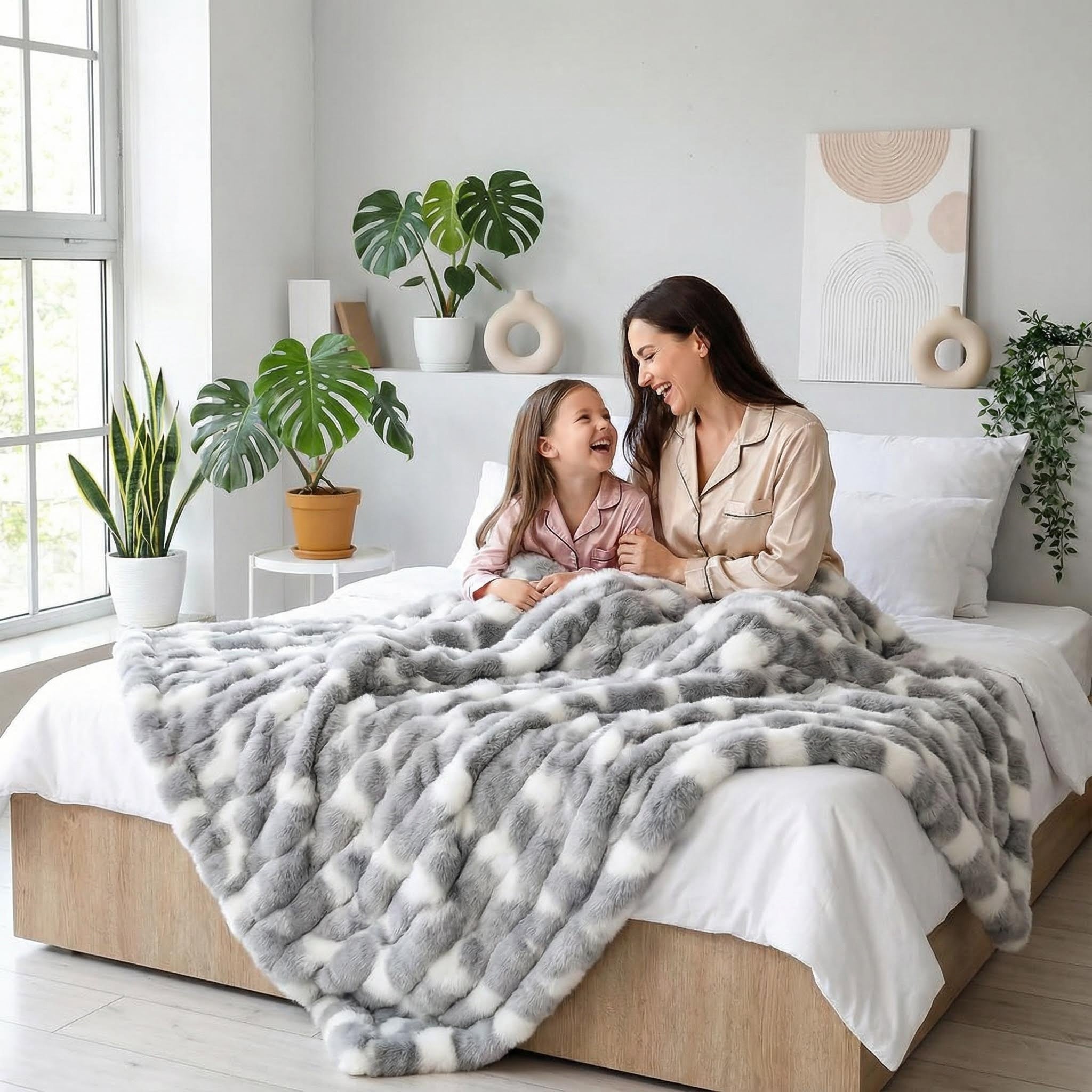 Mother and child laughing under a gray and white faux rabbit fur throw blanket on a bed.