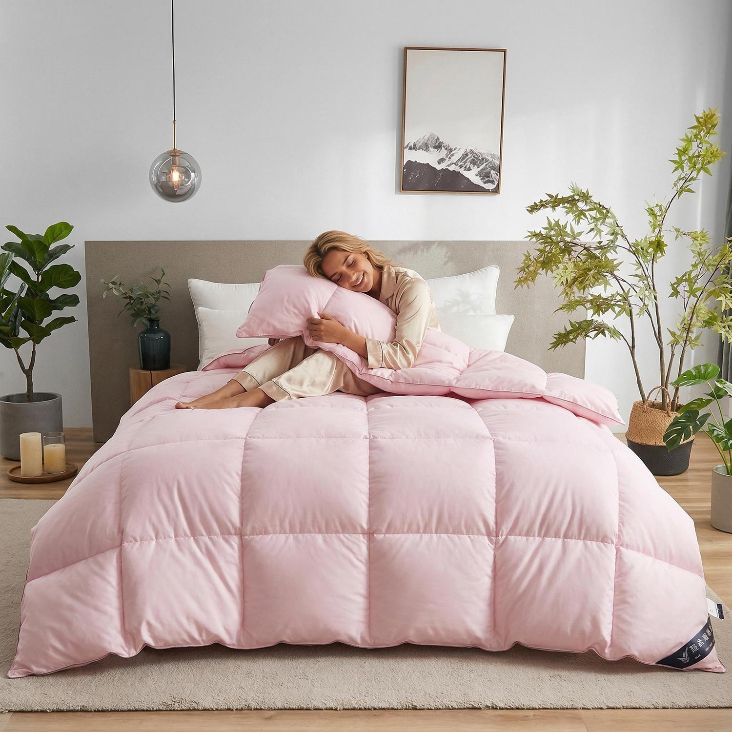 Smiling woman in pajamas on a bed with a soft pink goose down cotton quilt comforter and matching pillow.
