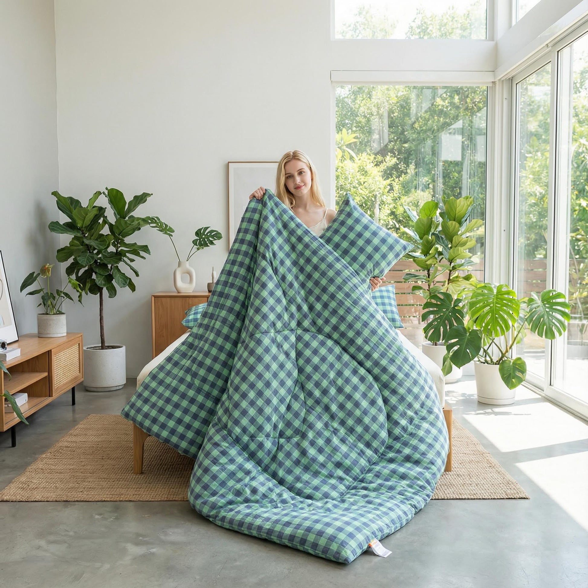 Blonde woman holding a green and blue gingham flannel fleece blanket over a bed in a bright room.