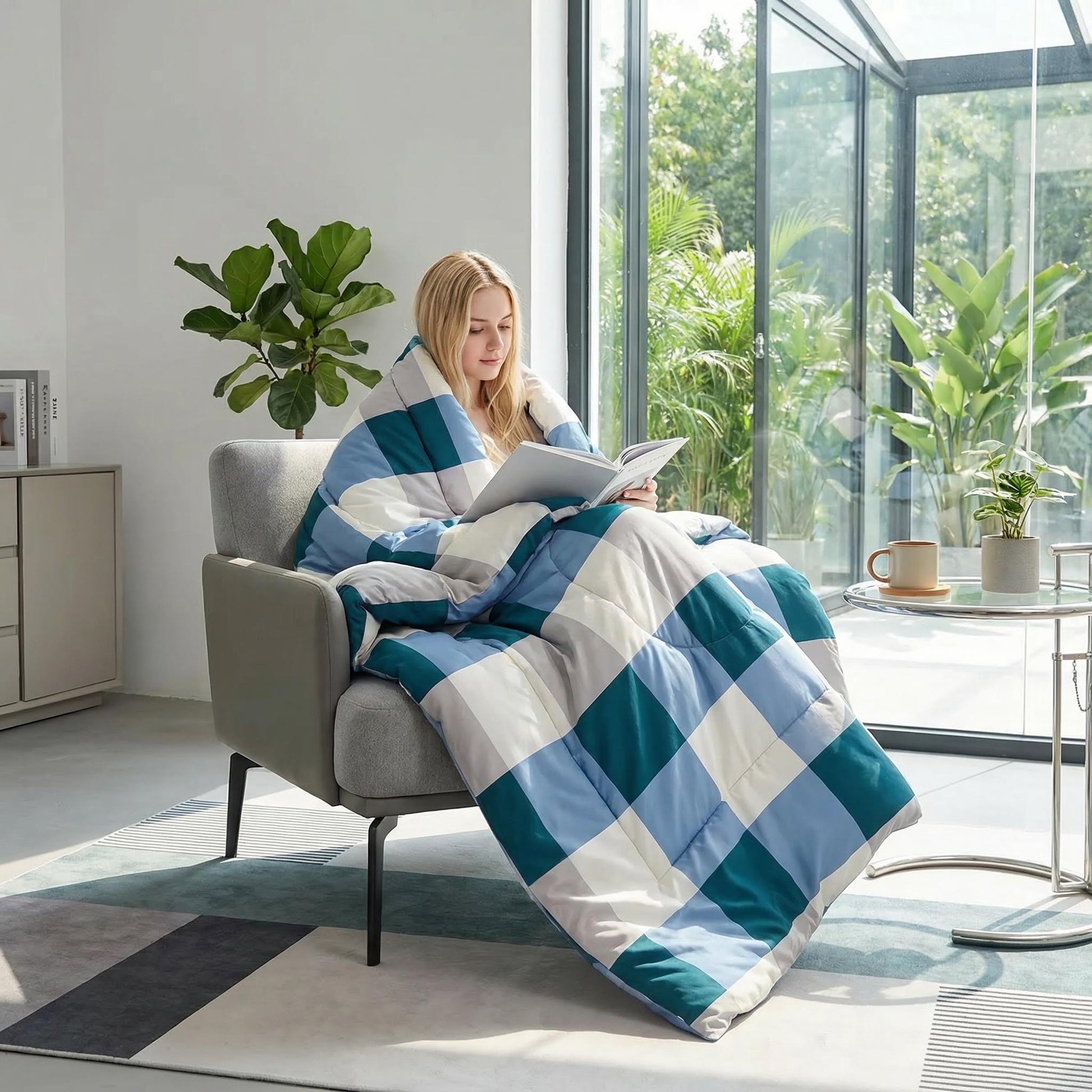 Blonde woman wrapped in a blue, teal, and white plaid flannel fleece blanket reading in a modern living room.