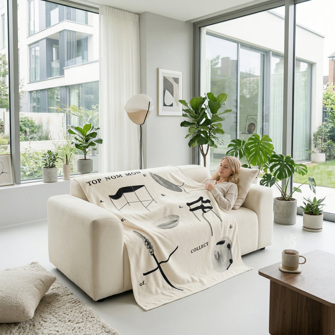Woman relaxing on a cream sofa under a white flannel throw blanket with black graphic designs in a modern living room.