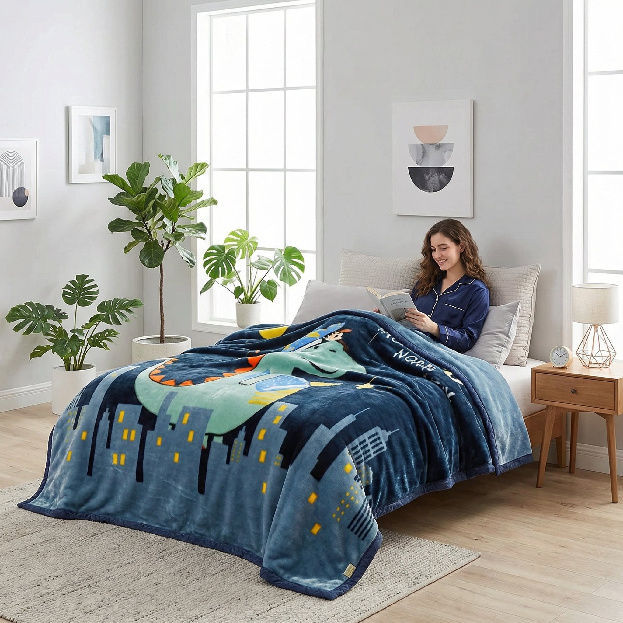 Woman reading in bed under a blue microfiber fleece throw blanket with a dinosaur and city design.