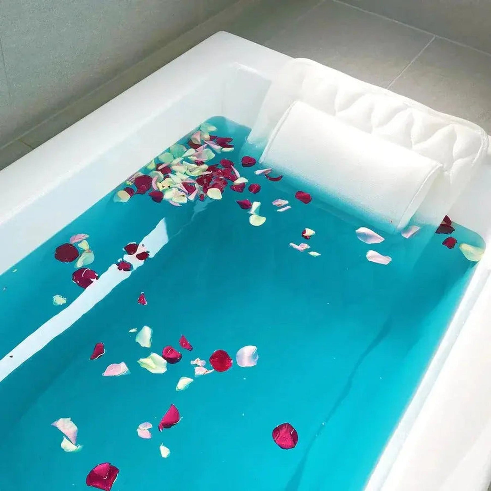 White mesh full body bath pillow with headrest in a tub filled with blue water and rose petals.