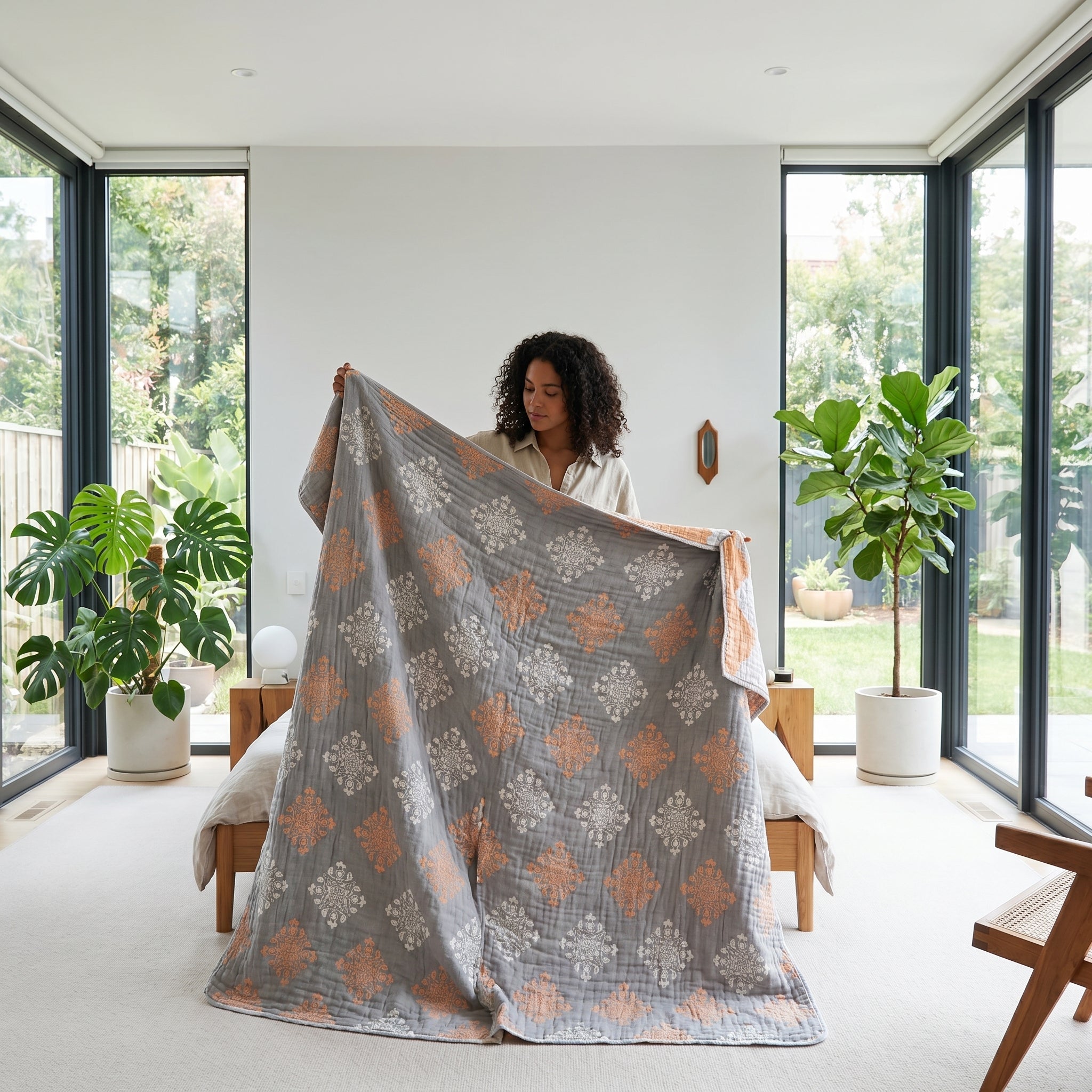 Woman holding a 6-layer cotton gauze blanket with a gray and peach reversible medallion pattern in a bright bedroom.