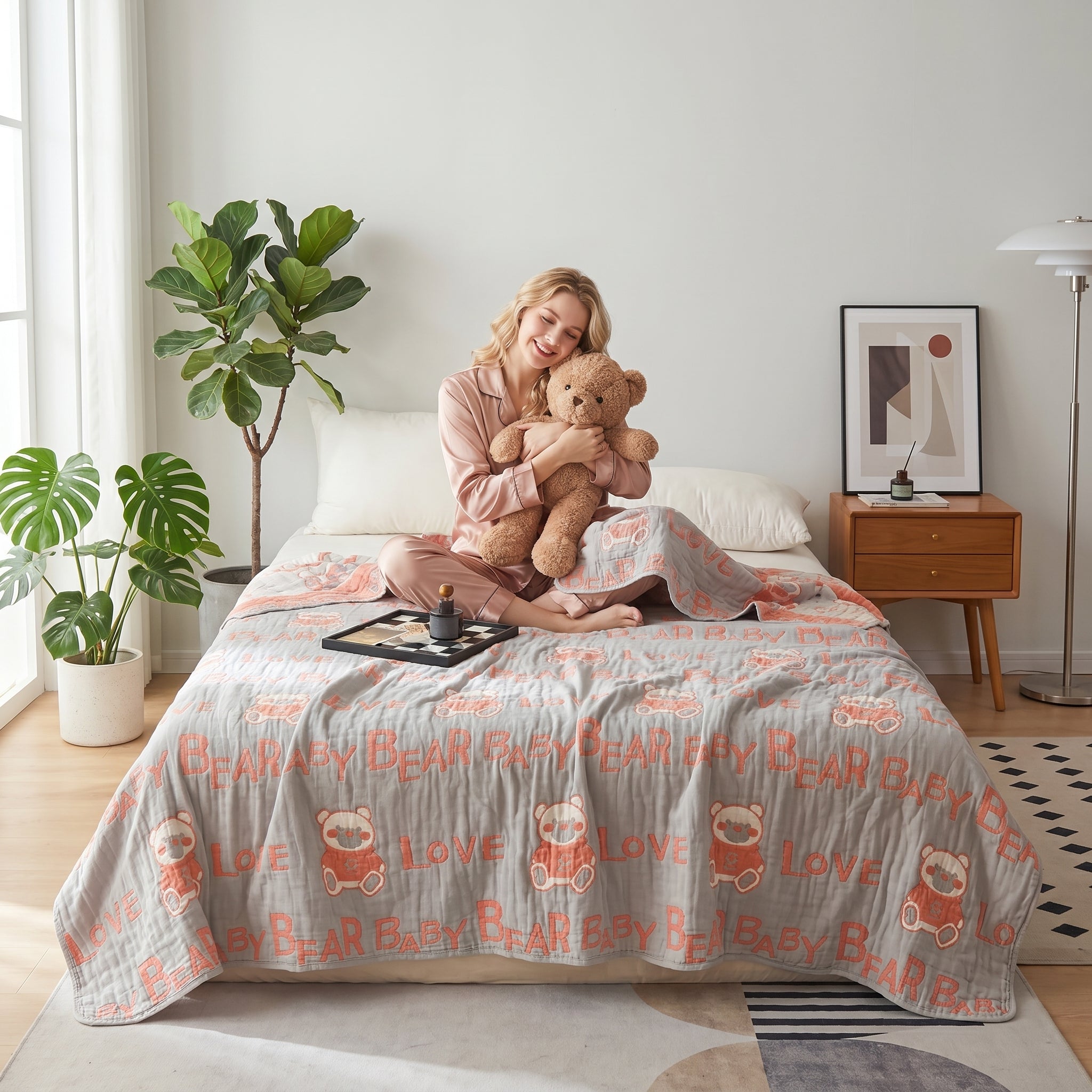 Smiling woman in pink pajamas on a bed with a gray 6-layer cotton gauze blanket featuring bear and "LOVE" print.