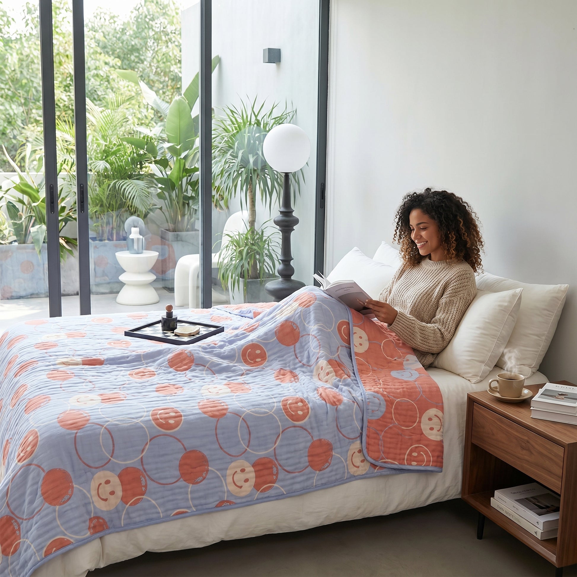 Woman reading in bed under a reversible 6-layer cotton gauze blanket with blue and peach smiley face patterns.