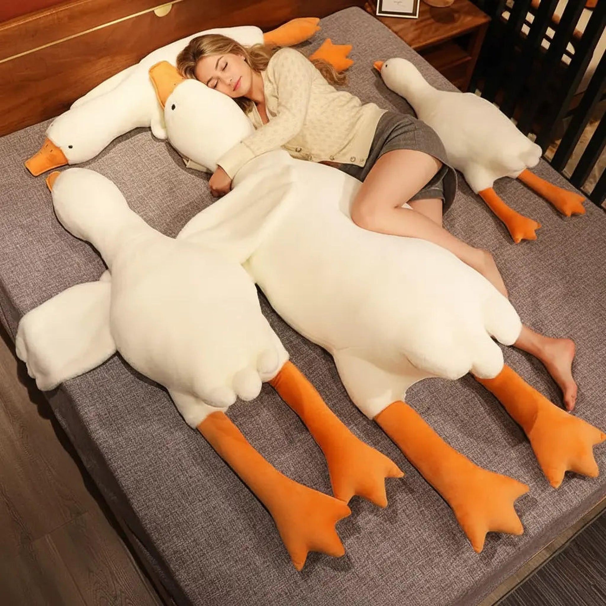 Woman sleeping on a bed hugged by giant white and orange goose plush pillow toys.