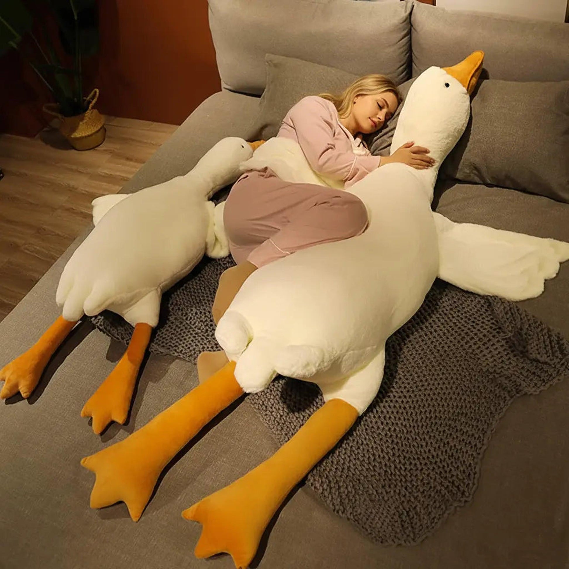 Woman sleeping in bed hugging a giant white goose plush pillow toy with orange legs.