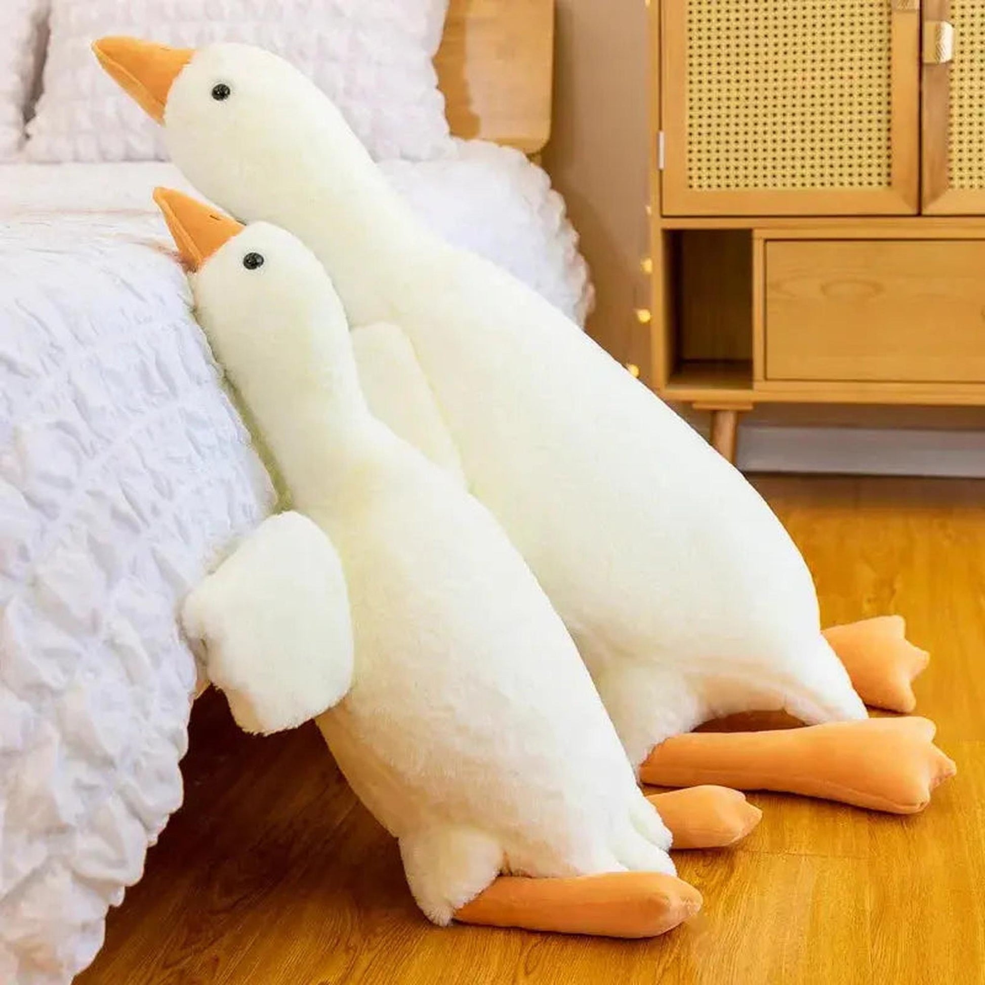 Two soft white goose plush pillow toys with orange beaks and feet leaning against a bed