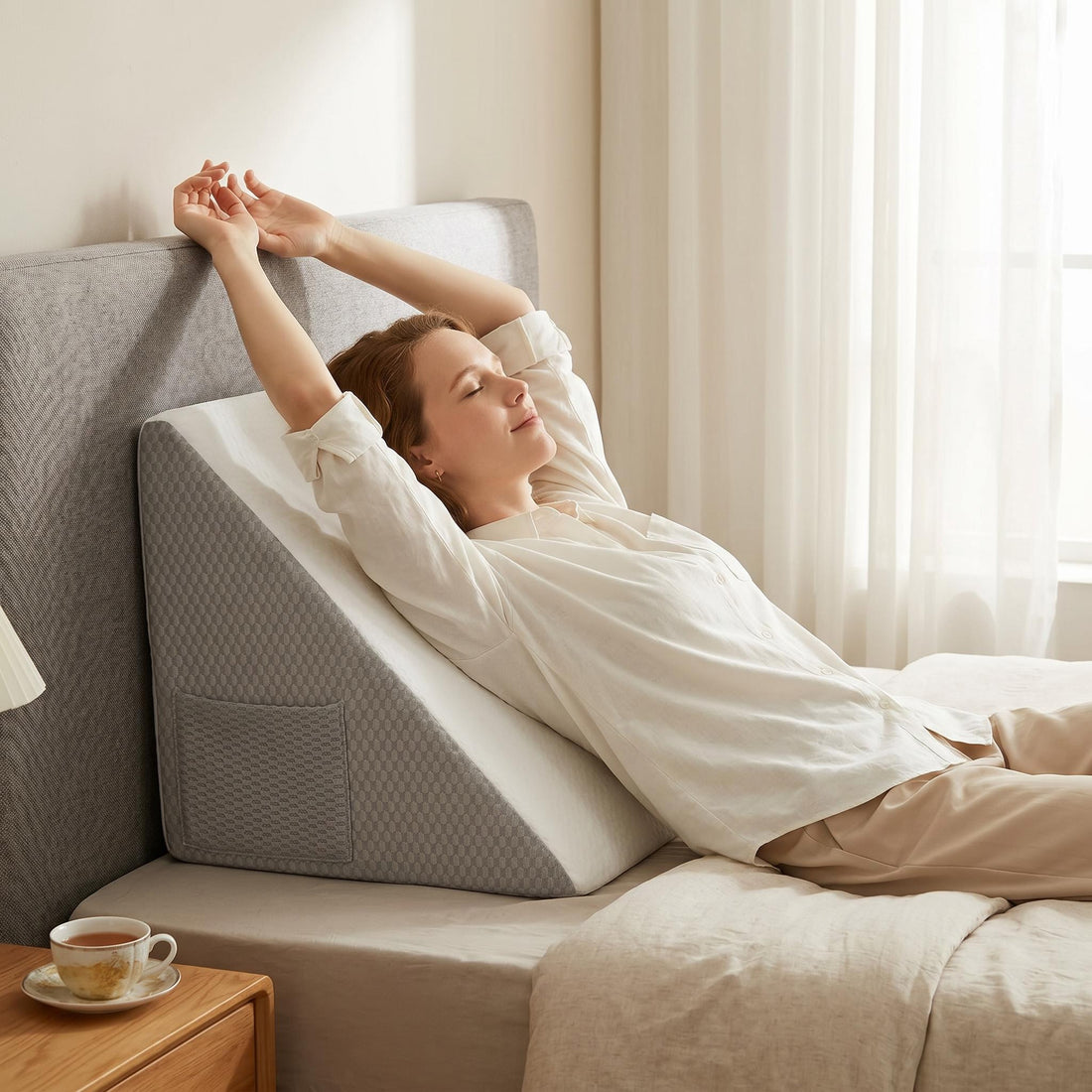 Woman using laptop in bed, supported by a grey and white inclined wedge pillow with side pockets.