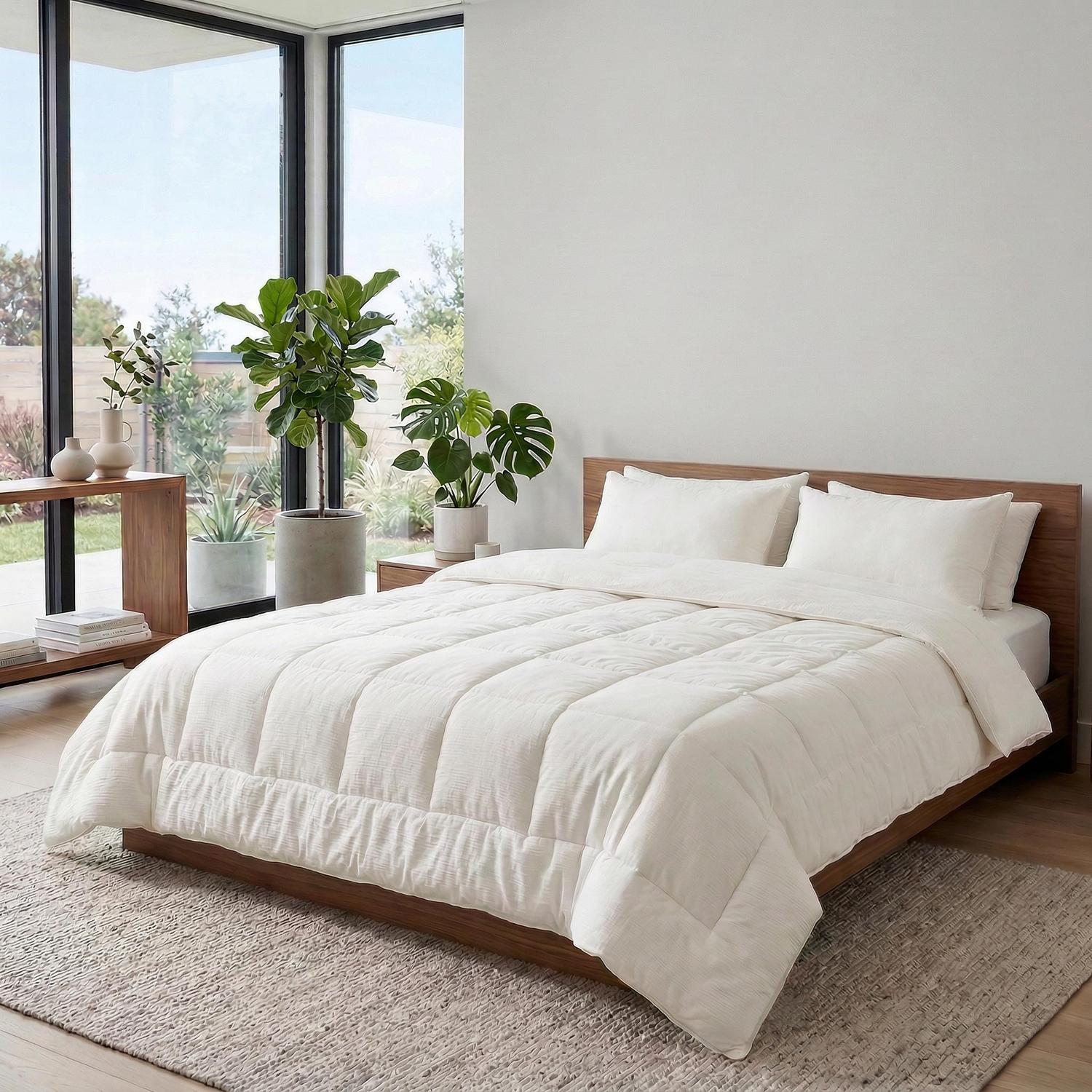 White quilted cotton comforter and two pillows on a wooden bed in a bright room.