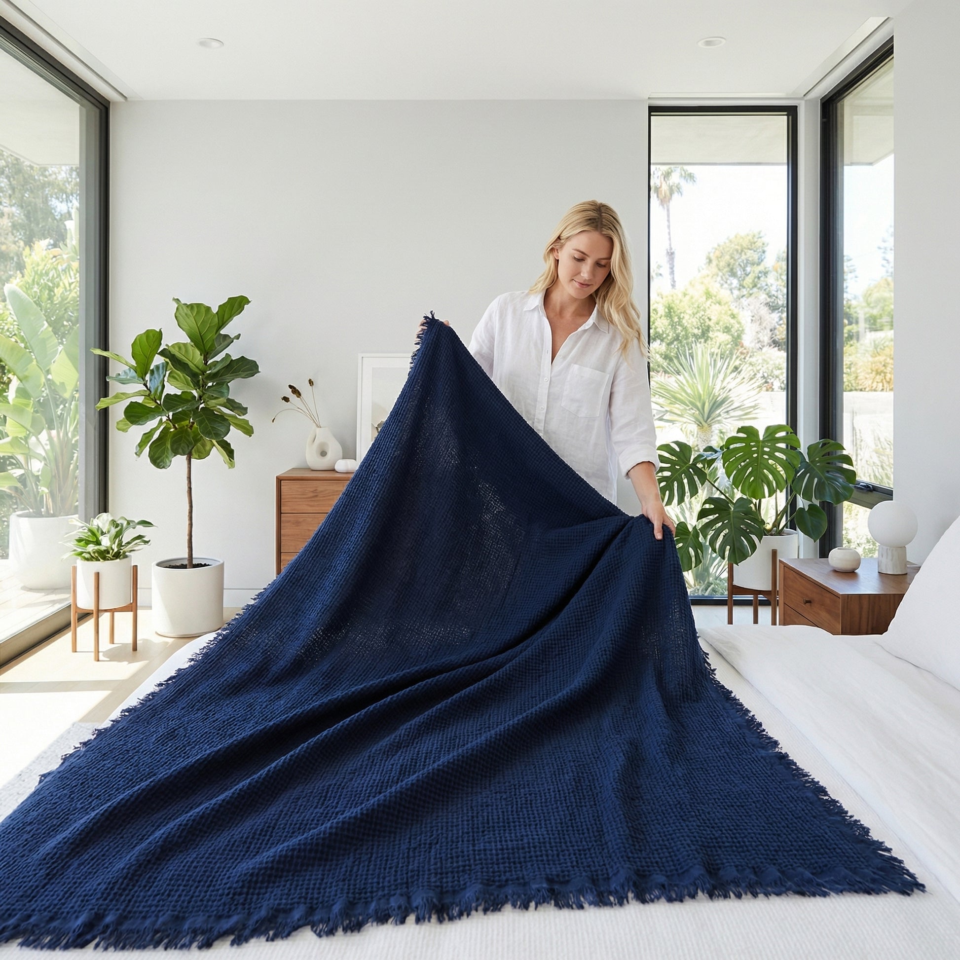 Woman placing a navy blue fringed cotton throw blanket on a white bed in a bright, modern room.