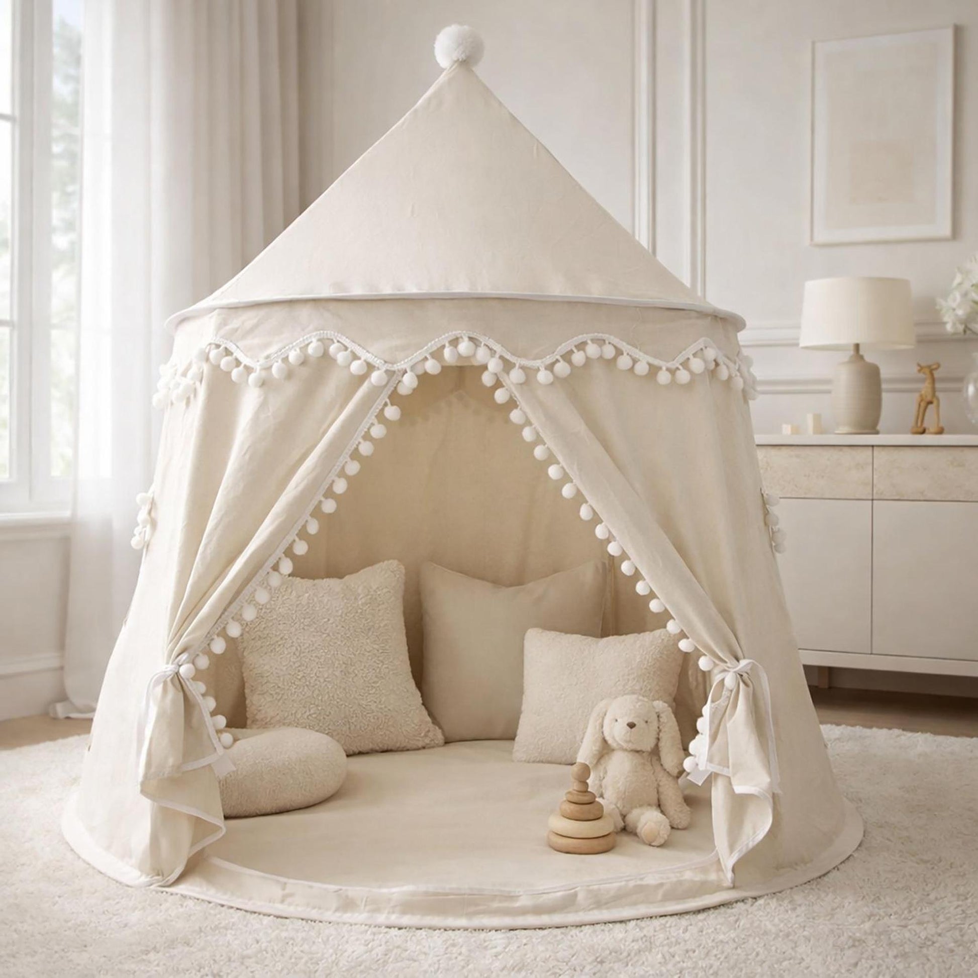 Cream fabric indoor play tent with pom-pom trim, tied-back opening, and cozy cushions on a rug.