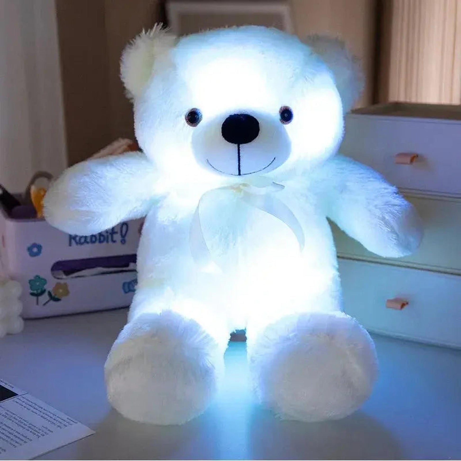 White plush teddy bear glowing brightly with color-changing LED lights, featuring a small white bow.