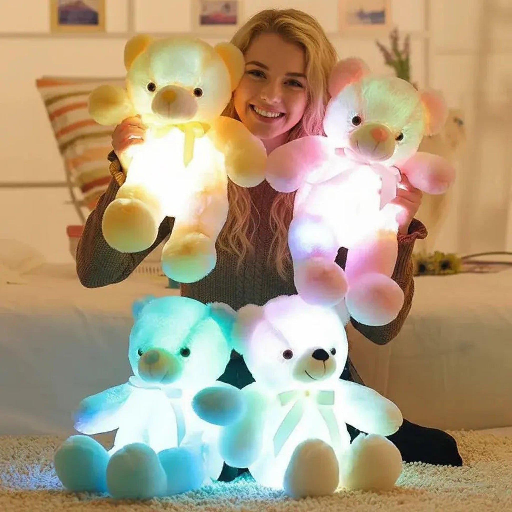 Smiling woman holding two glowing plush teddy bears, one yellow and one pink, with two more blue and white bears on the floor.