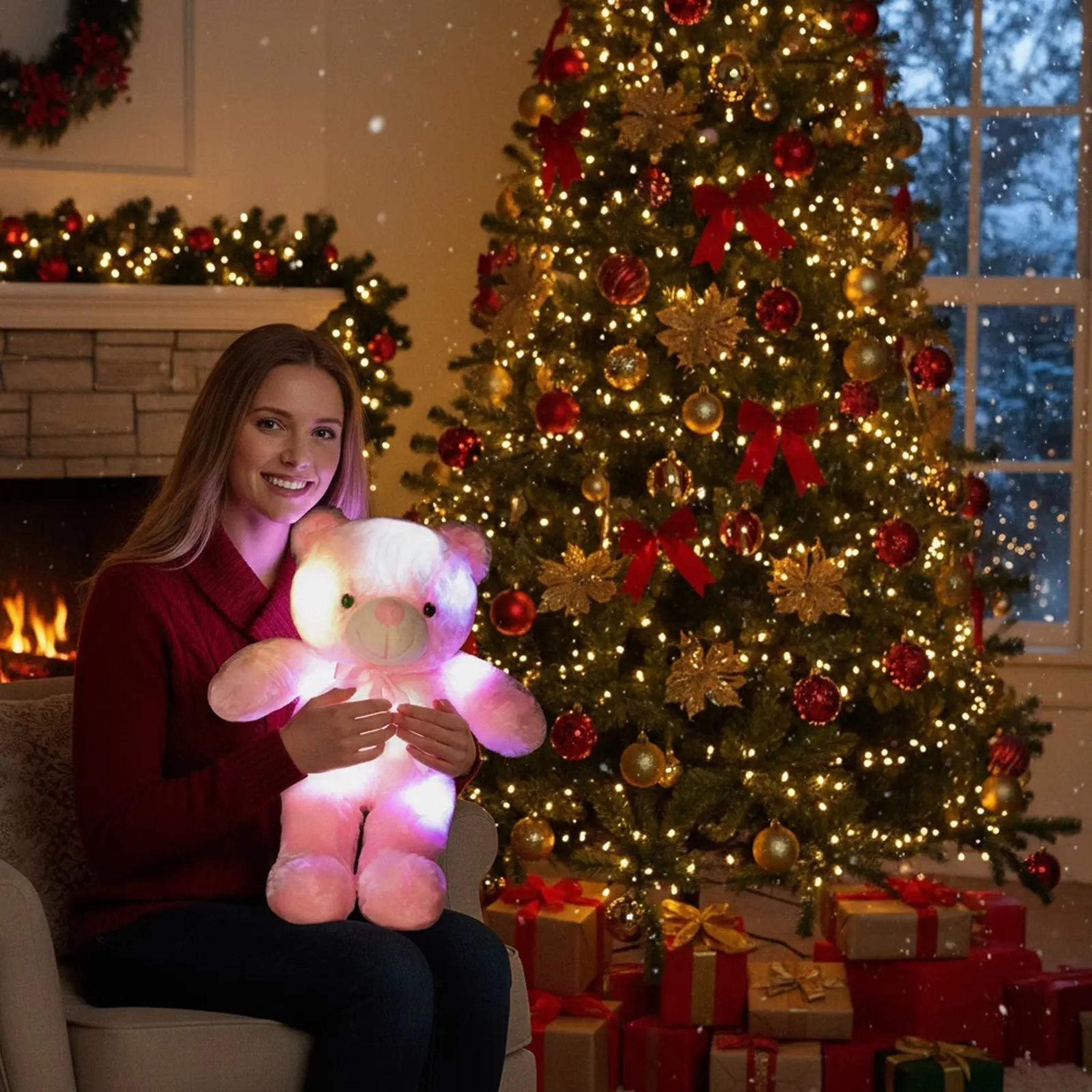 Smiling woman holding a glowing pink plush teddy bear in front of a lit Christmas tree and fireplace.