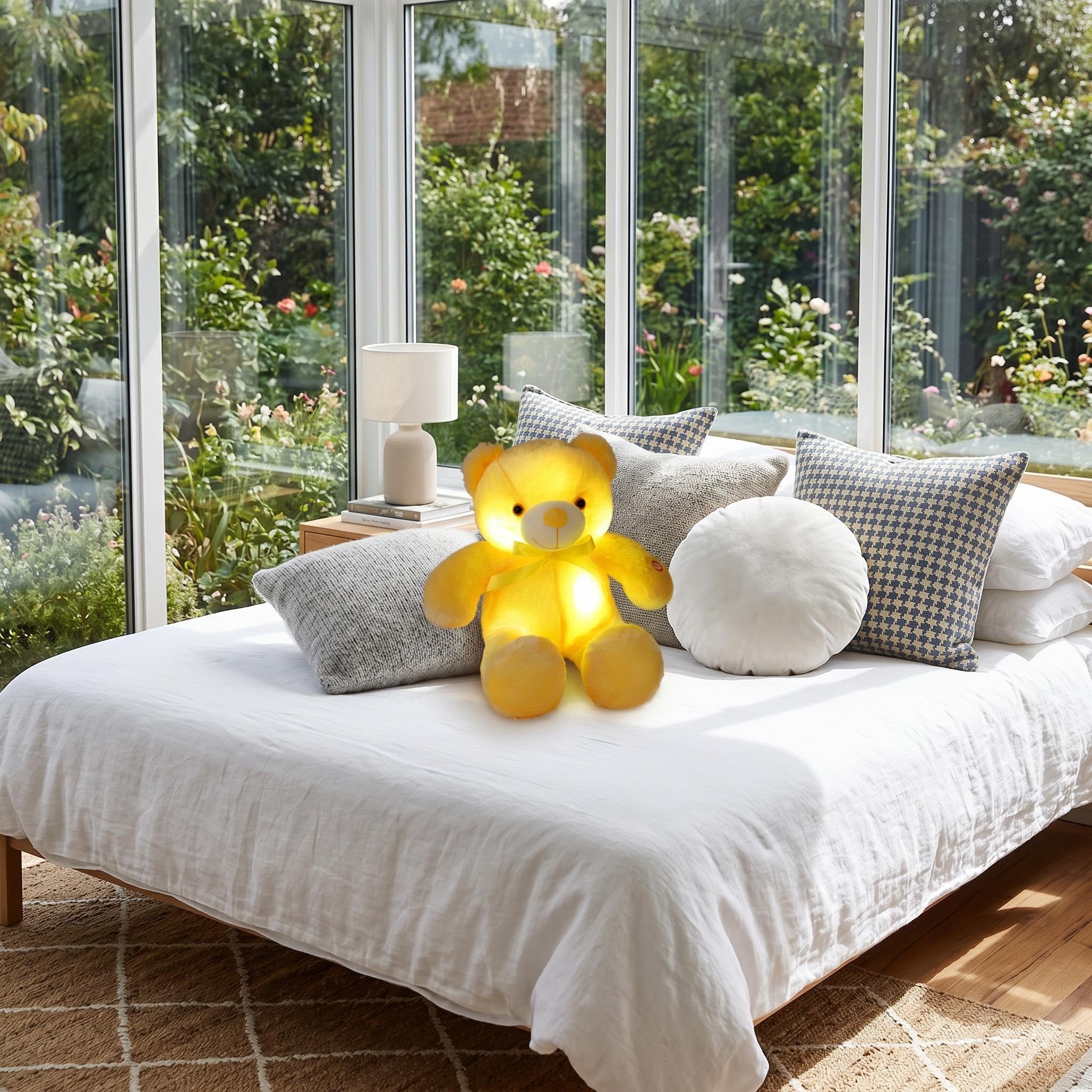 Glowing yellow plush teddy bear with color-changing LED light, sitting on a white bed in a bright room.