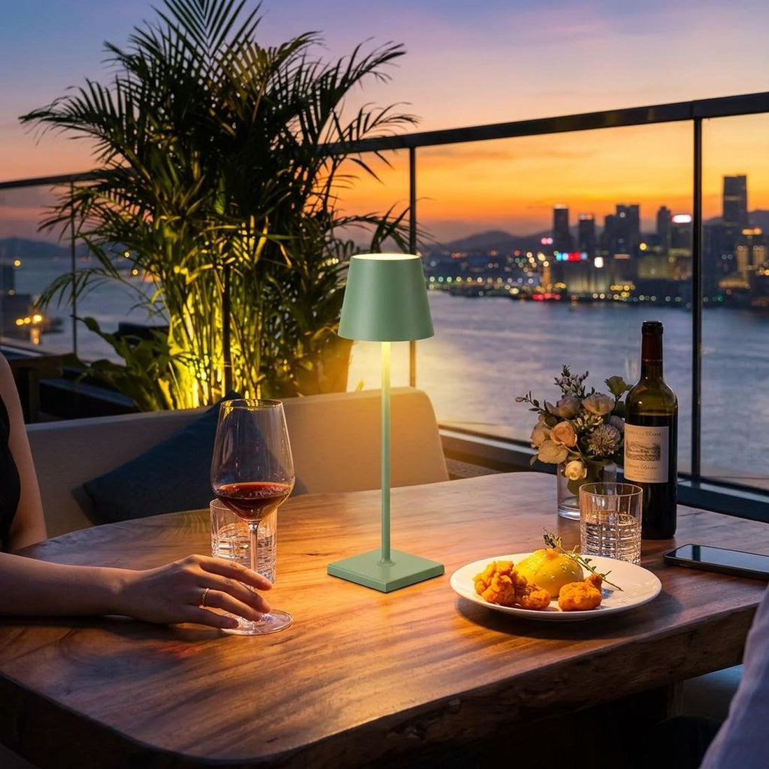 Sage green portable LED table lamp with a minimalist aluminum design on an outdoor dining table at sunset.