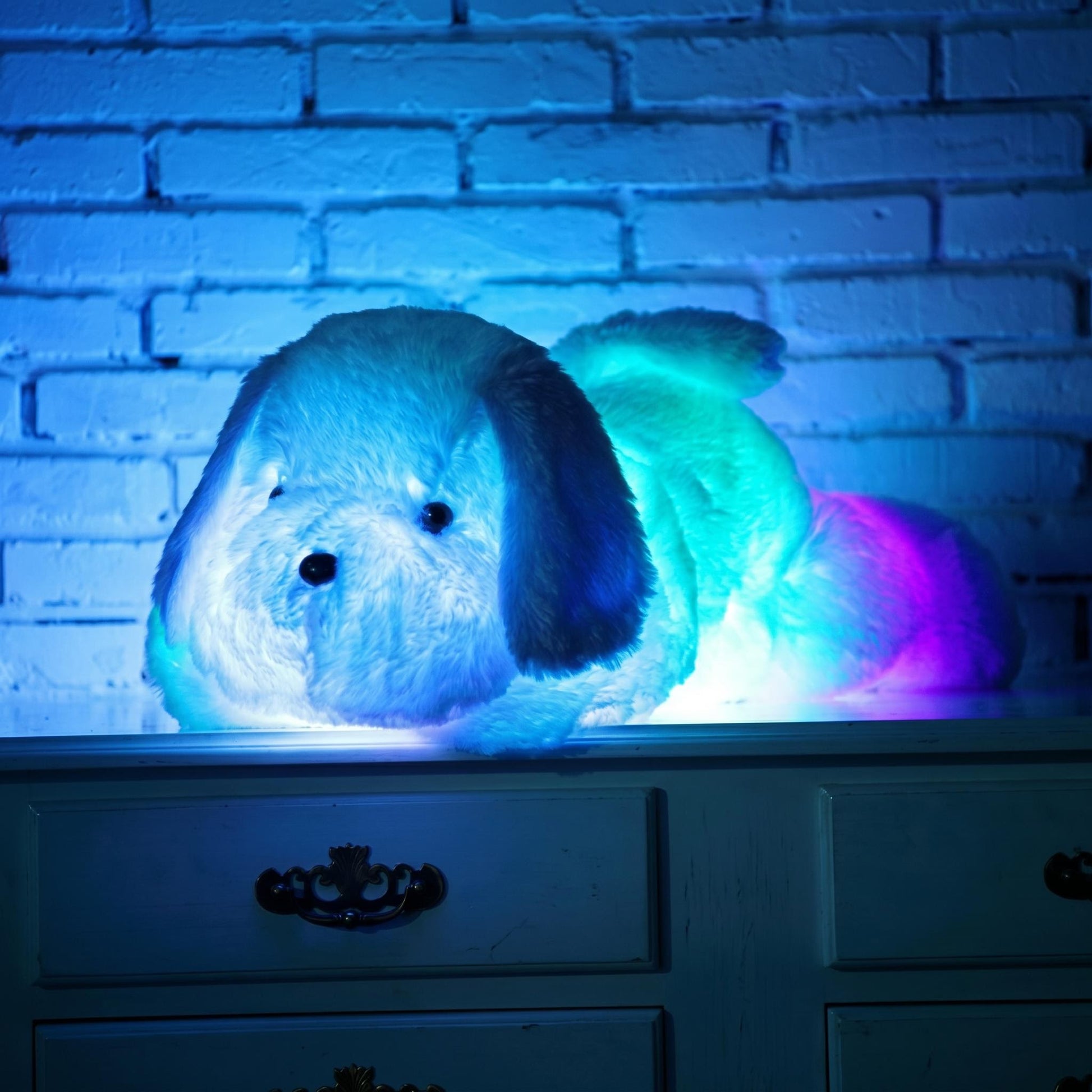 Luminous dog plush pillow glowing with colorful LED lights on a dresser against a brick wall.