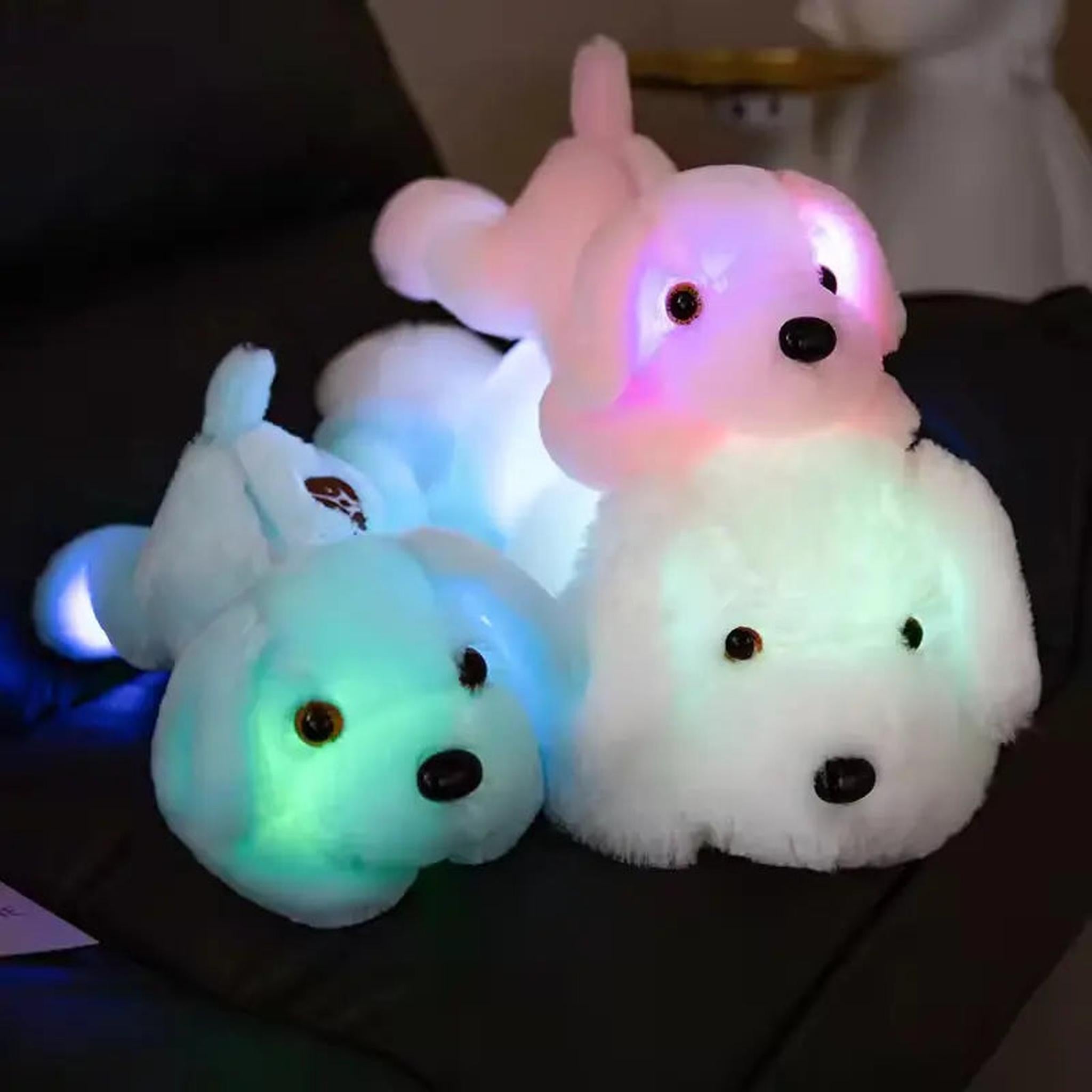 Three luminous dog plush pillows glowing in pink, blue, and white LED lights on a dark sofa.