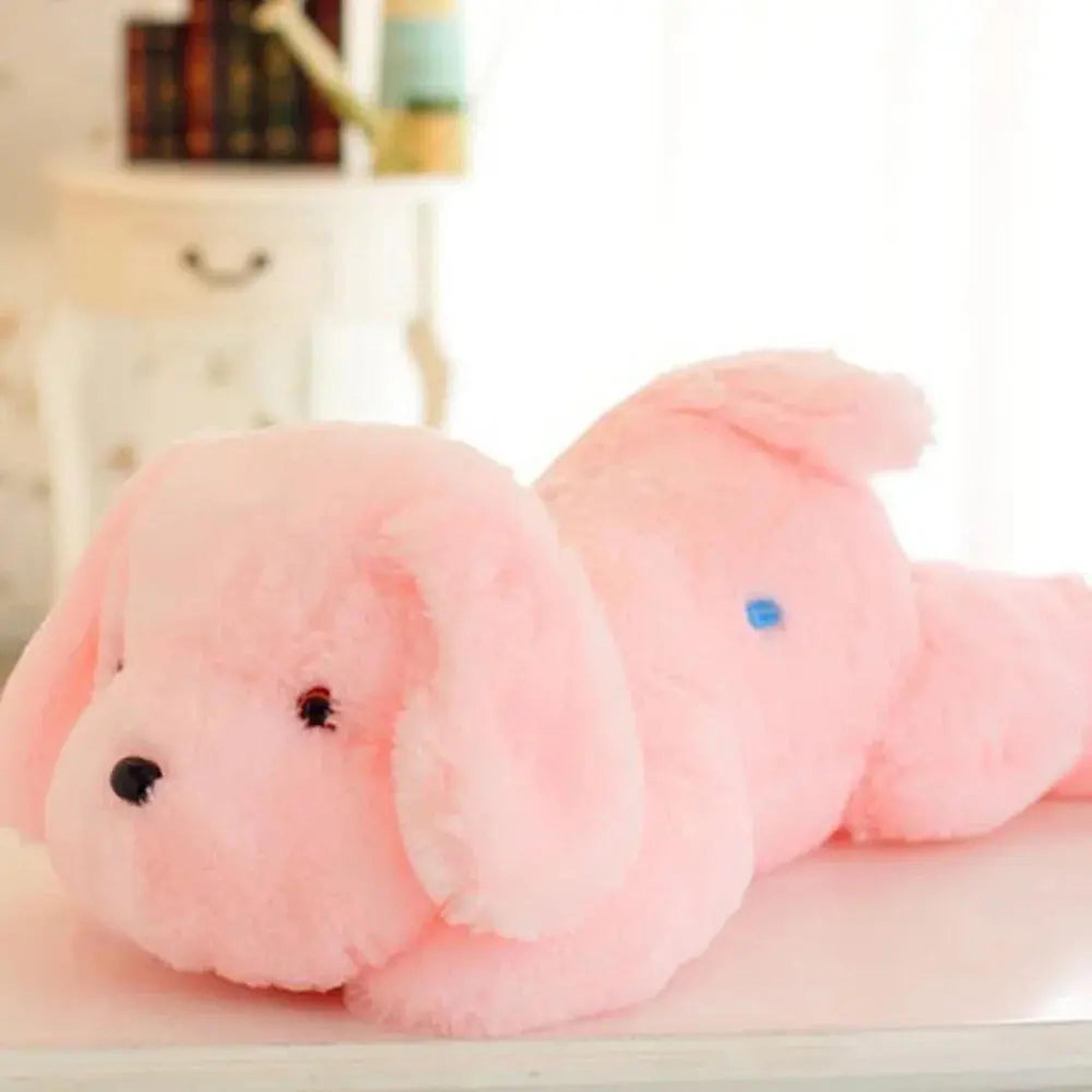 Soft pink plush dog pillow with LED light glowing blue on its back, lying on a white surface.