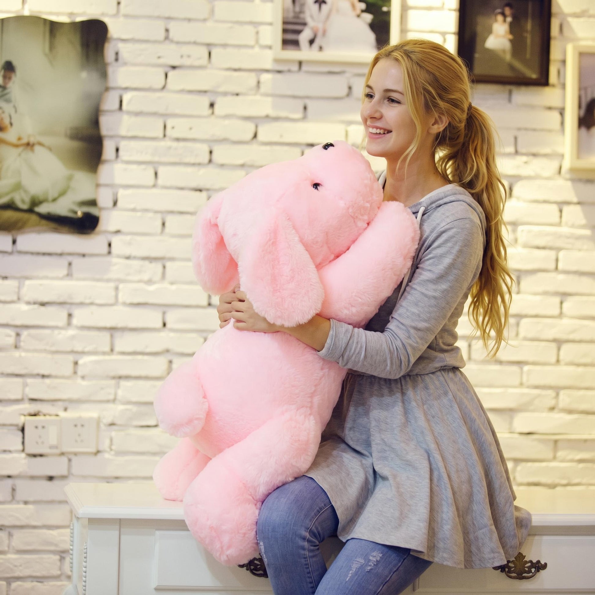 A smiling woman hugs a large, soft pink plush dog pillow in front of a white brick wall.