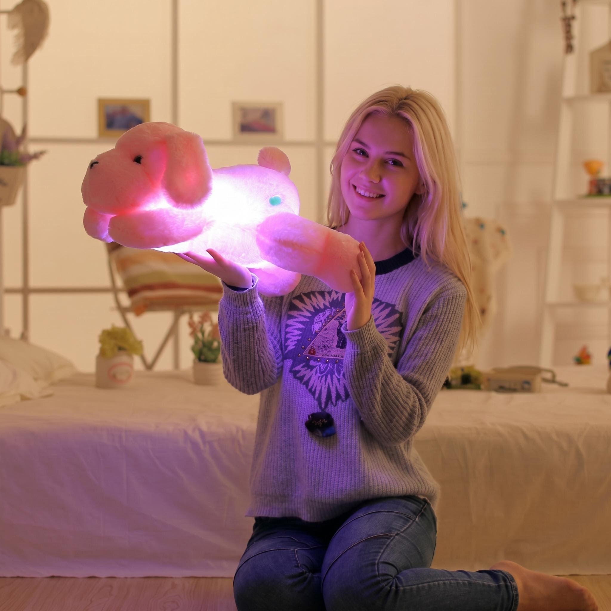 Smiling woman holding a glowing pink luminous dog plush pillow with LED light in a bedroom.