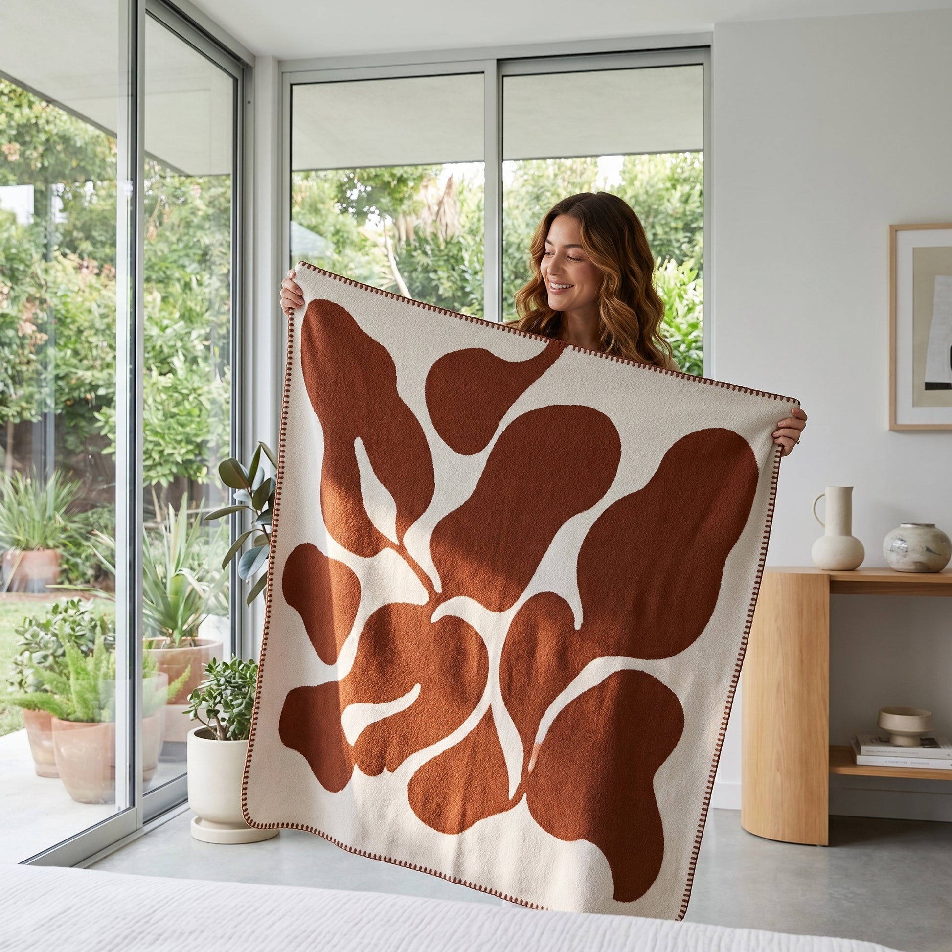 Woman holding a soft woven throw blanket with a brown abstract pattern on a cream background.