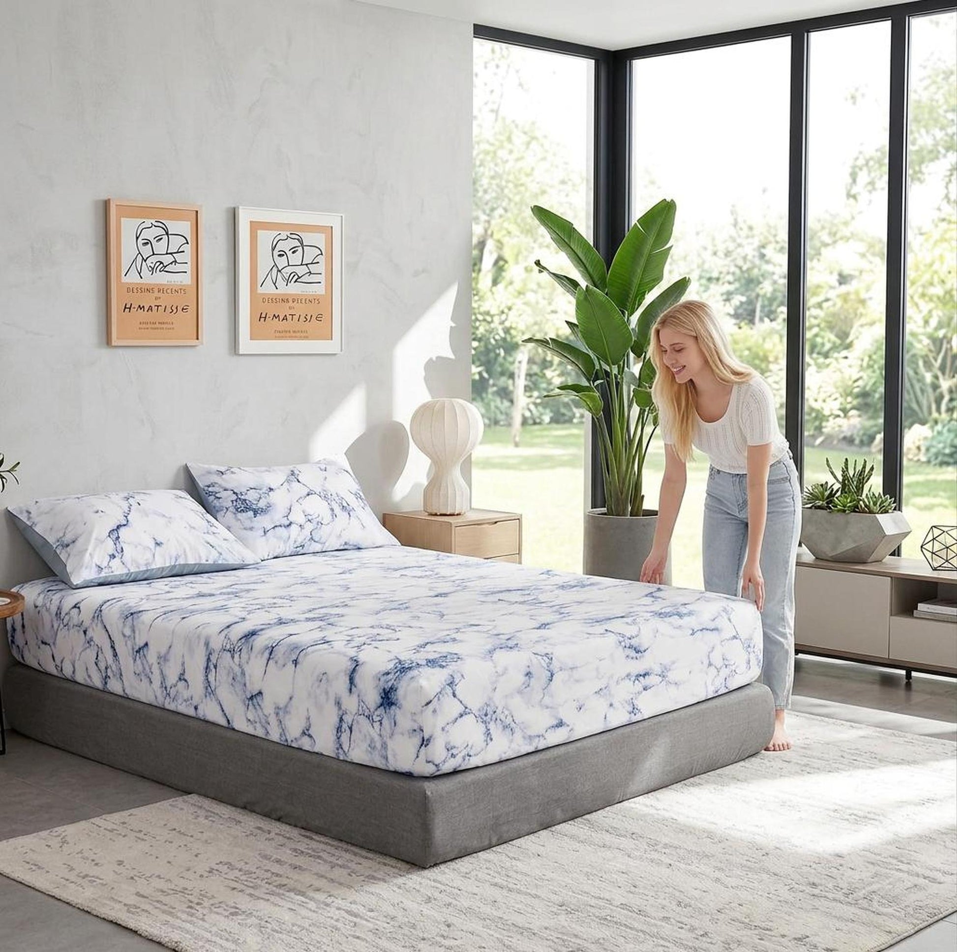 Woman adjusting a white and blue marble print microfiber fitted sheet on a grey bed in a modern bedroom.