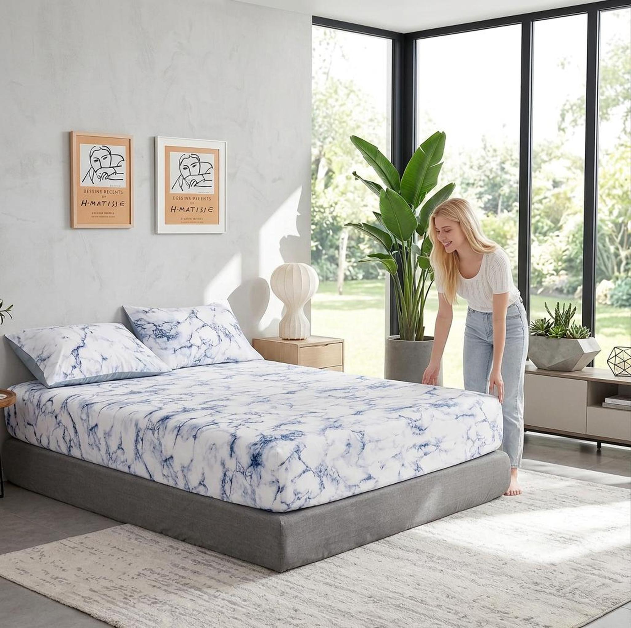 Woman adjusting a white and blue marble print microfiber fitted sheet on a grey bed in a modern bedroom.