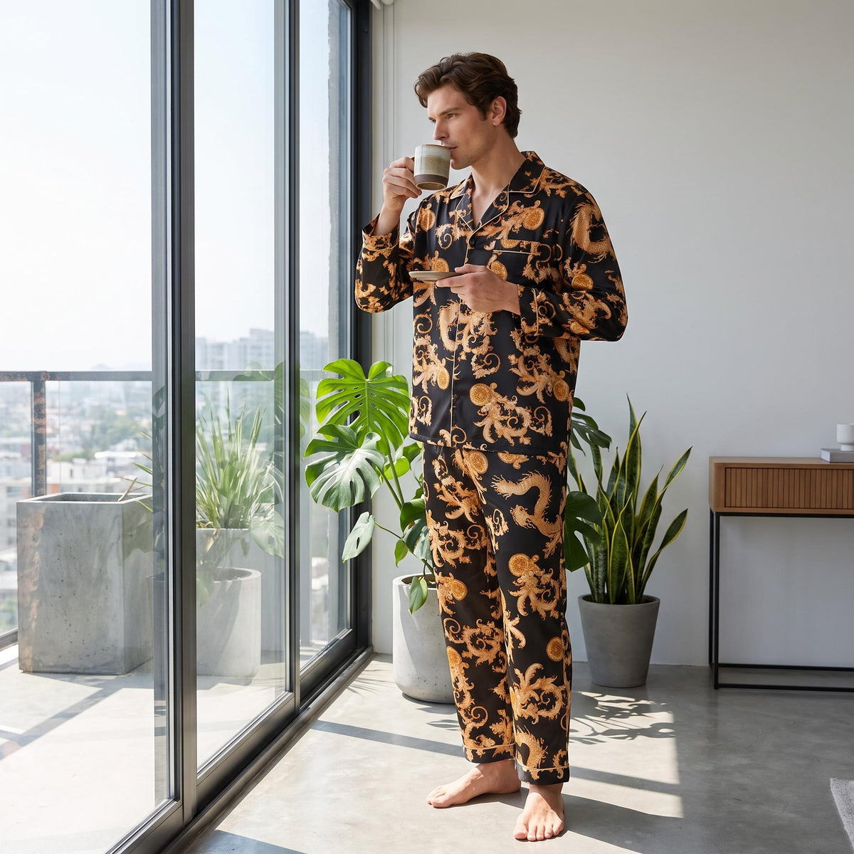 Man wearing a black and gold dragon print silk pajama set with a notch collar, standing by a window with a coffee cup.