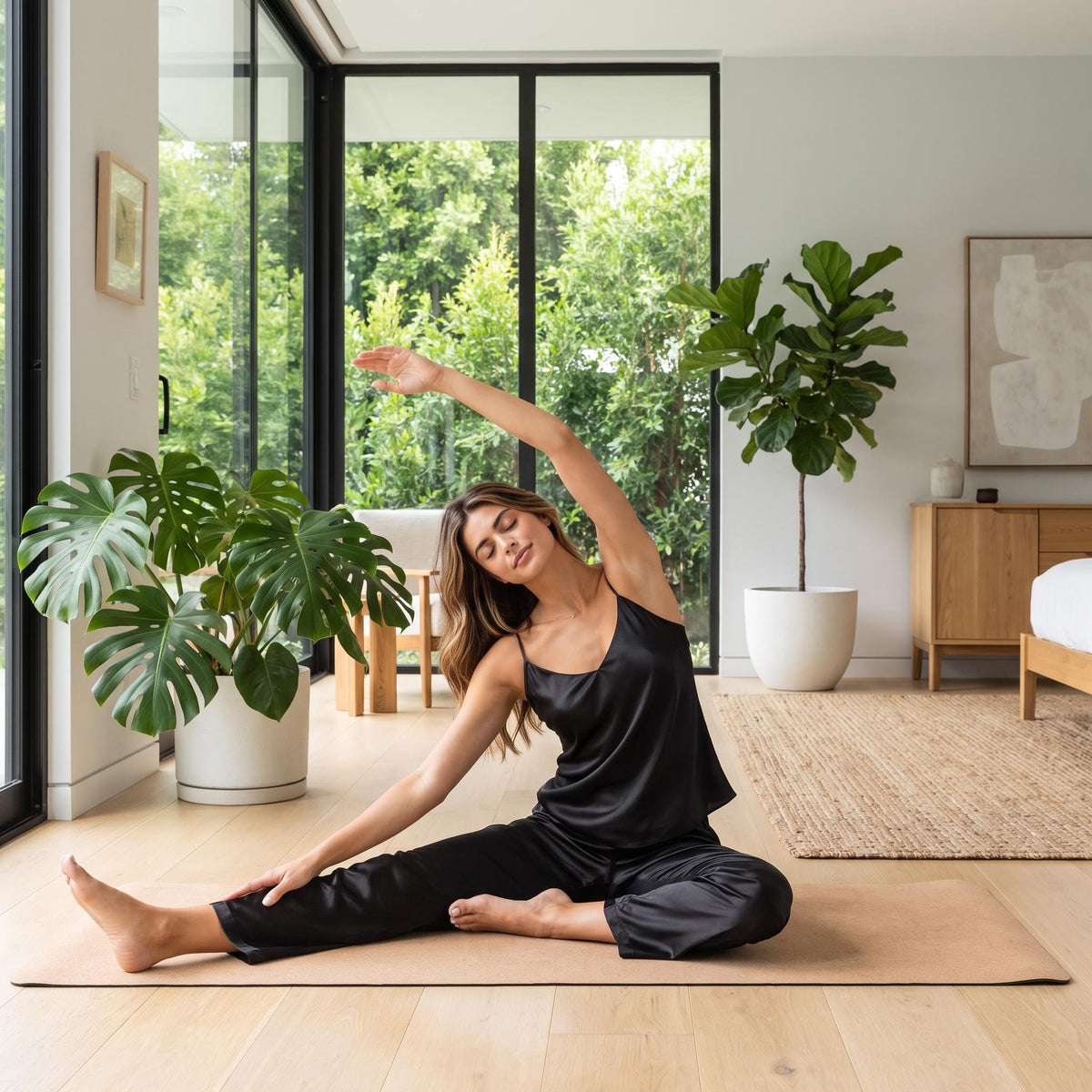 Woman wearing a black mulberry silk V-neck camisole and straight-leg pajama pants stretching on a yoga mat.