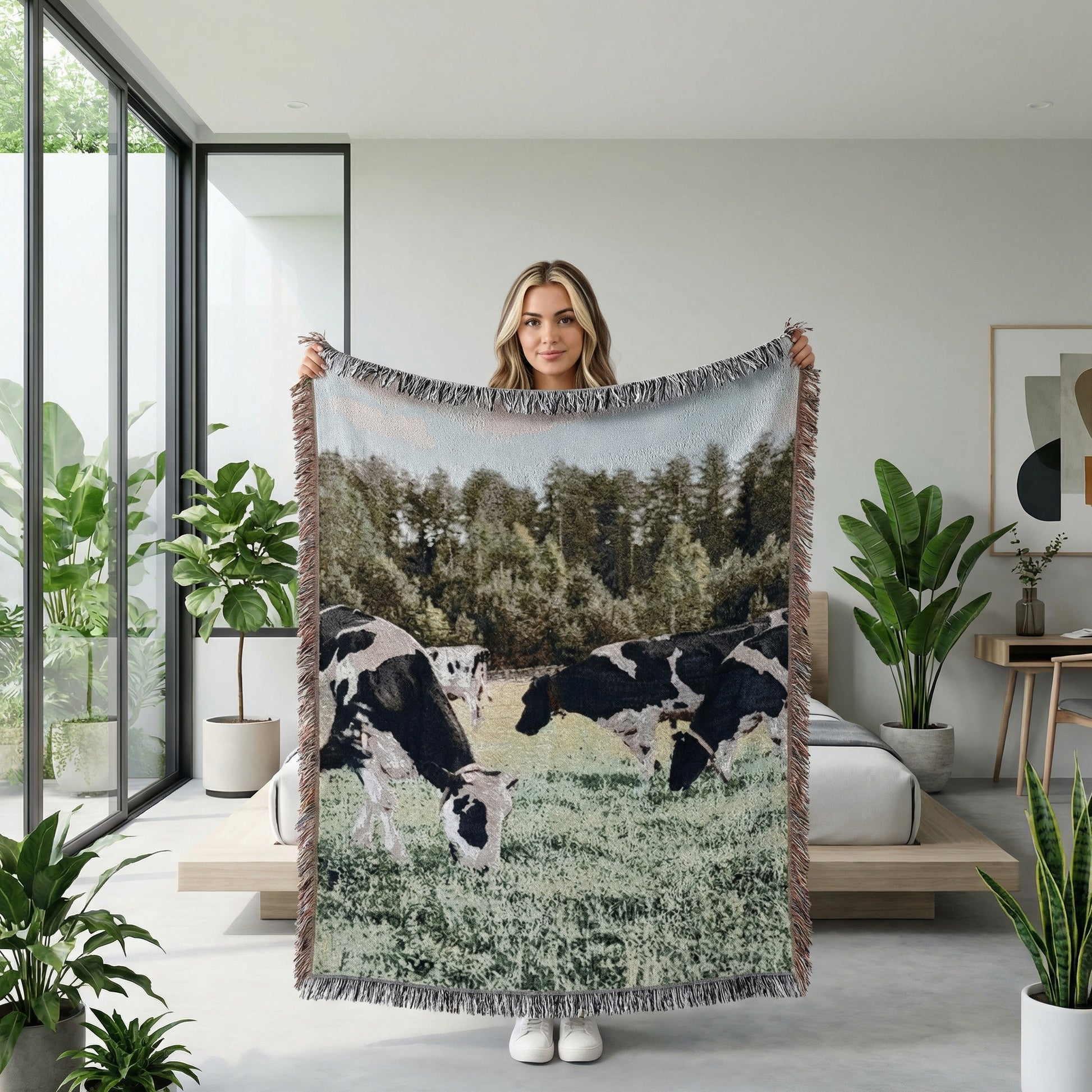 Woman holding a large knitted blanket with a natural landscape design of black and white cows grazing.