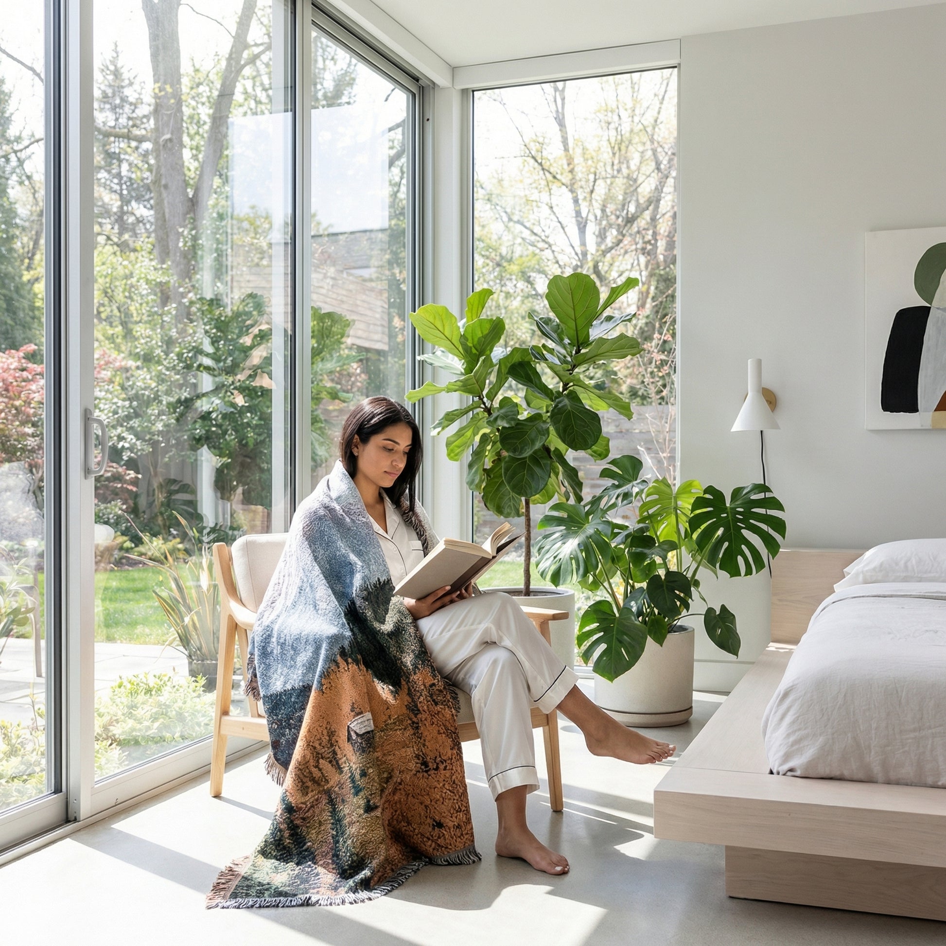 Woman reading in a bright room, wrapped in a large natural landscape knitted blanket with forest and sky design.